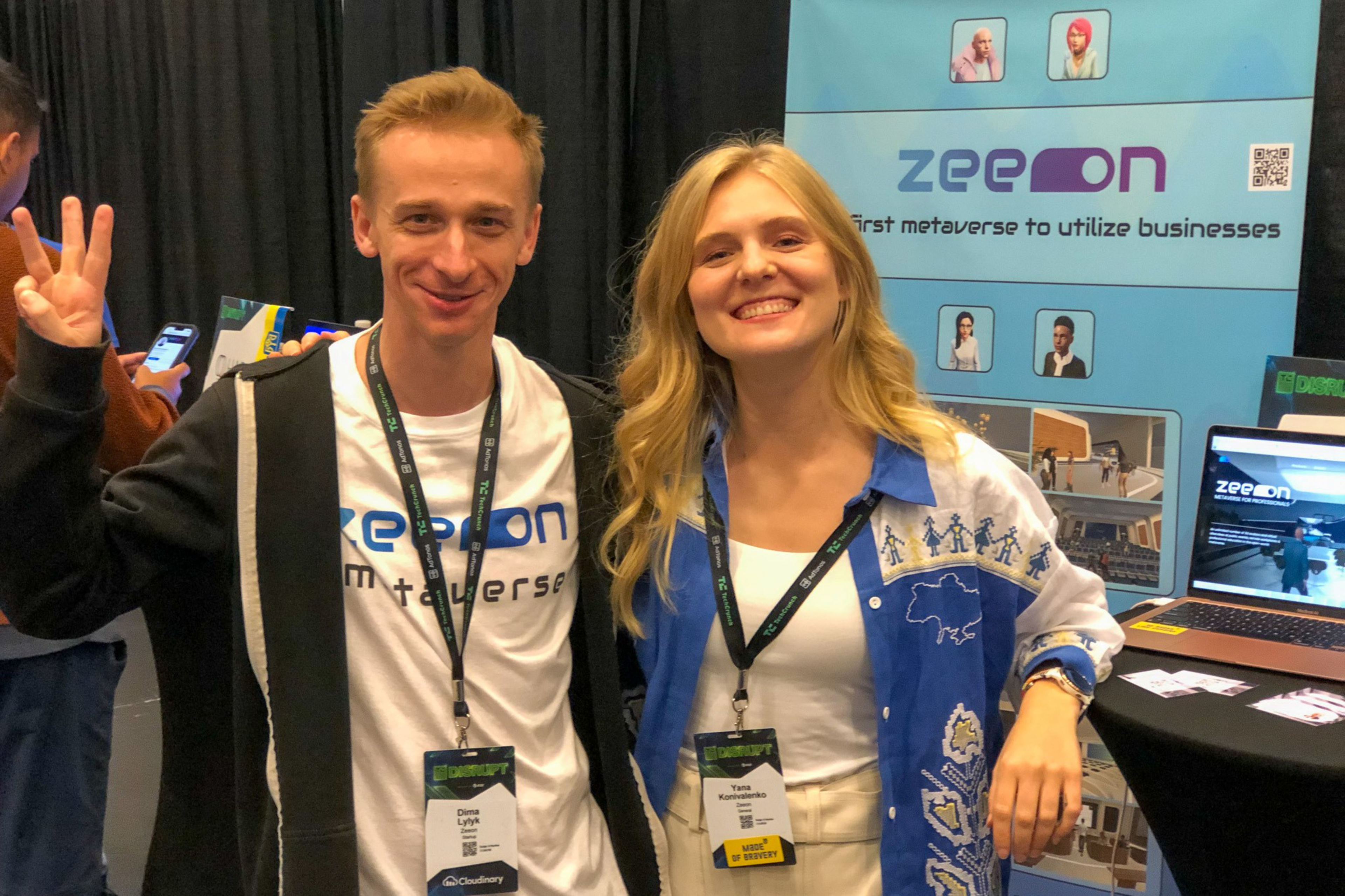 Two smiling people wear badges and stand in front of a booth with a blue banner that reads "zeeson first metaverse to utilize businesses."