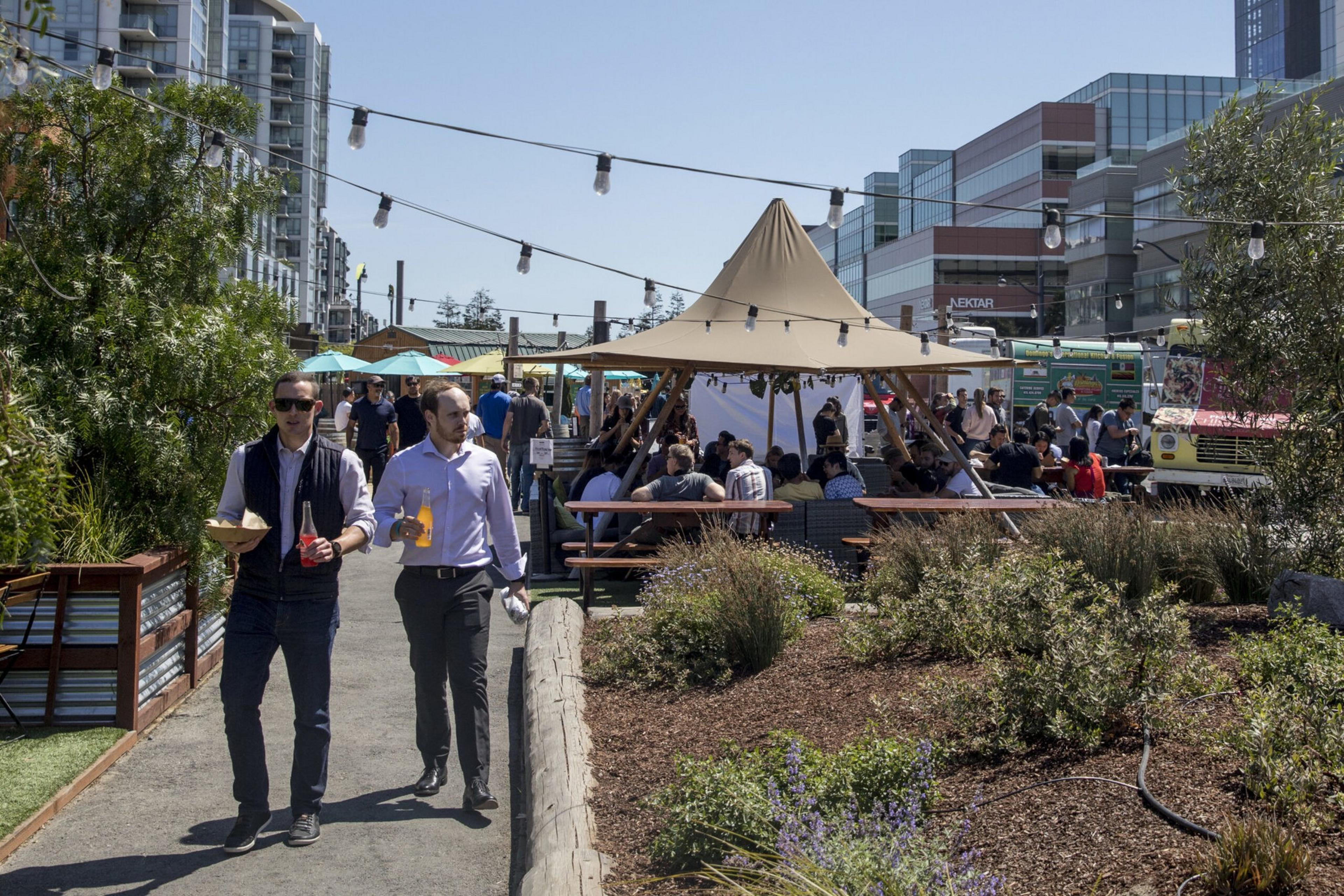 Two men walk on a pathway holding food and drinks near a shaded outdoor seating area, surrounded by plants and city buildings in the background.