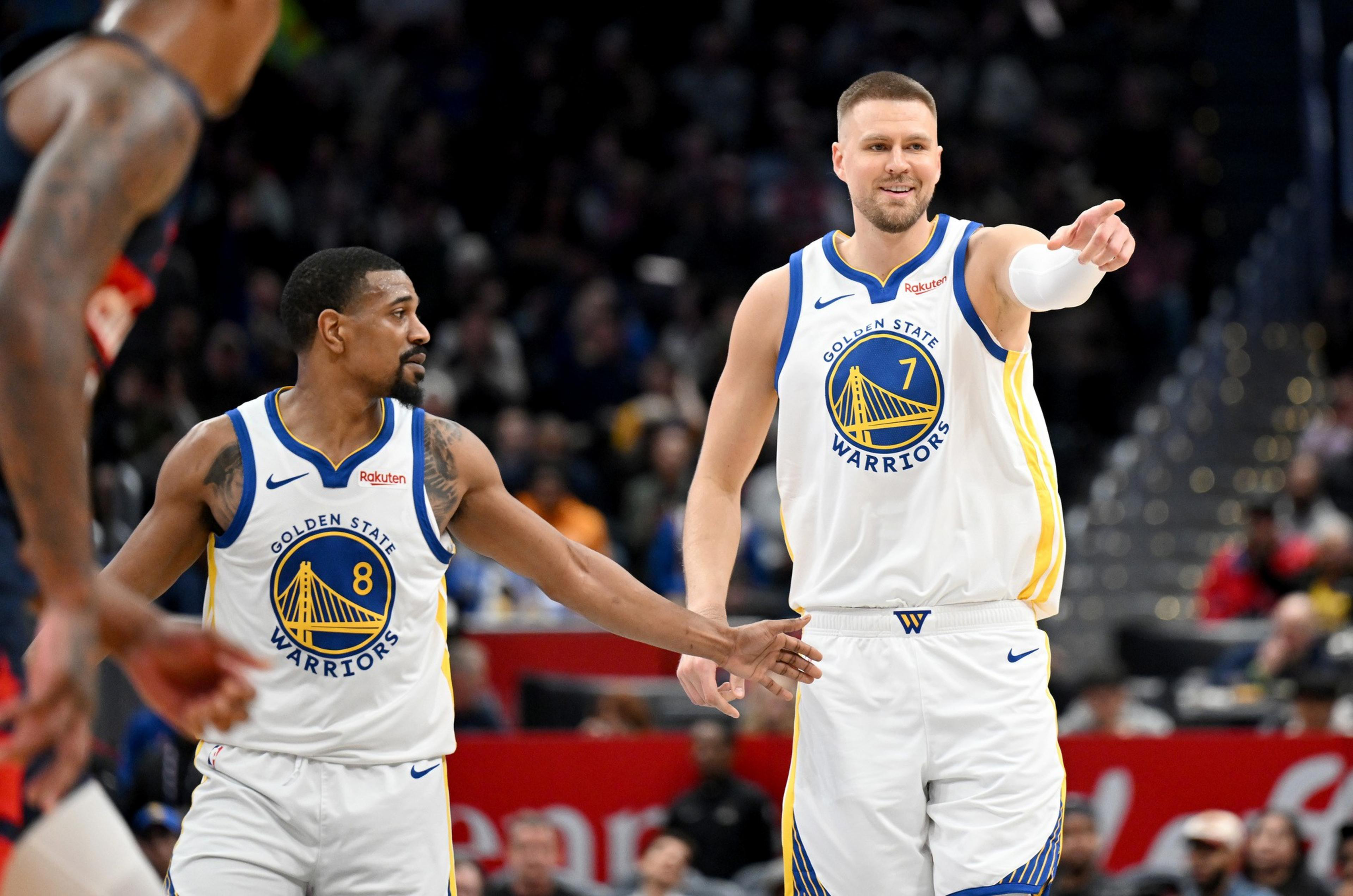 Two Golden State Warriors players in white jerseys interact on the basketball court, with one pointing and the other reaching out, while a blurred opponent stands nearby.
