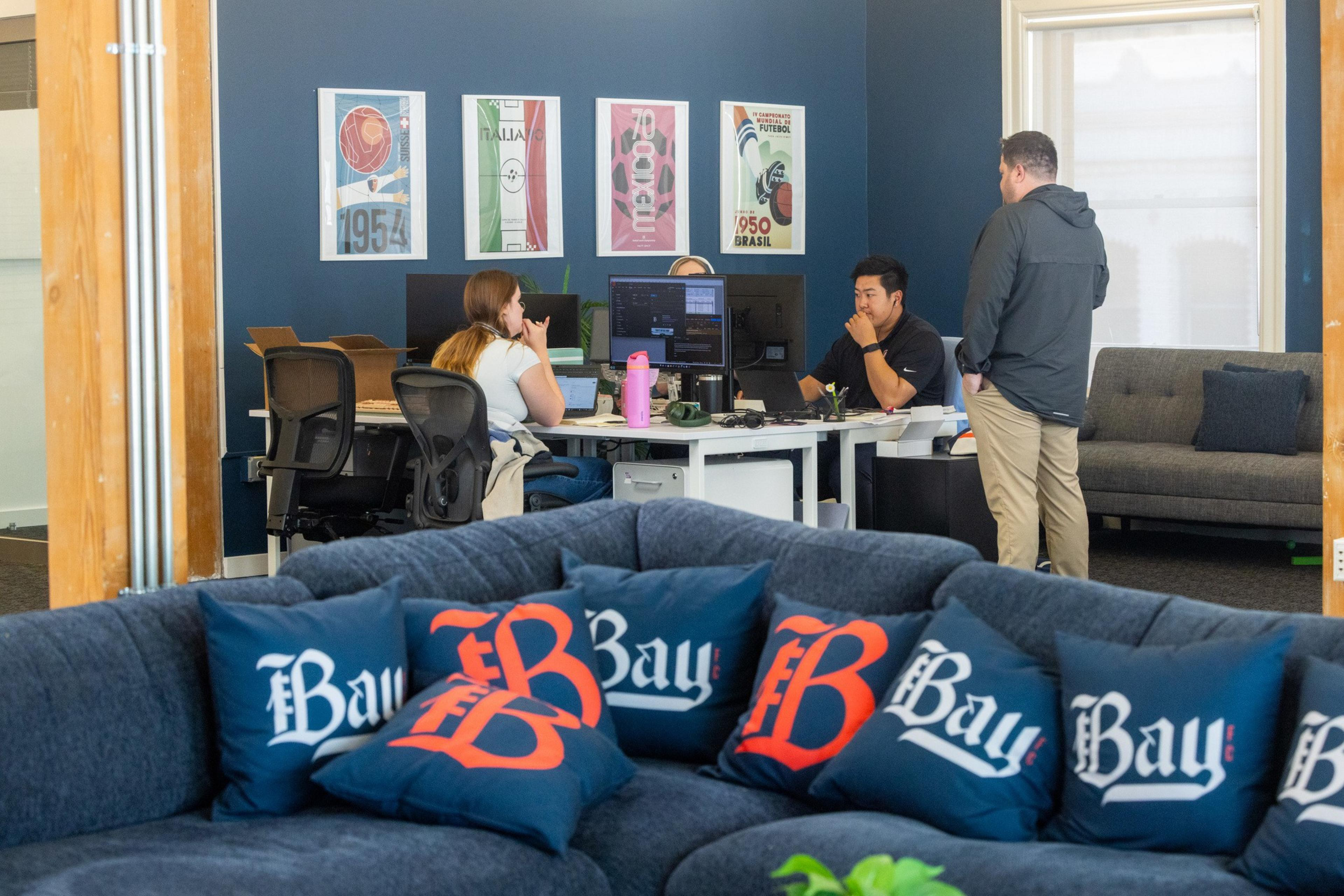 Three people are working and discussing at desks in an office with a blue wall and framed sports posters; the foreground features a couch with blue "Bay" pillows.