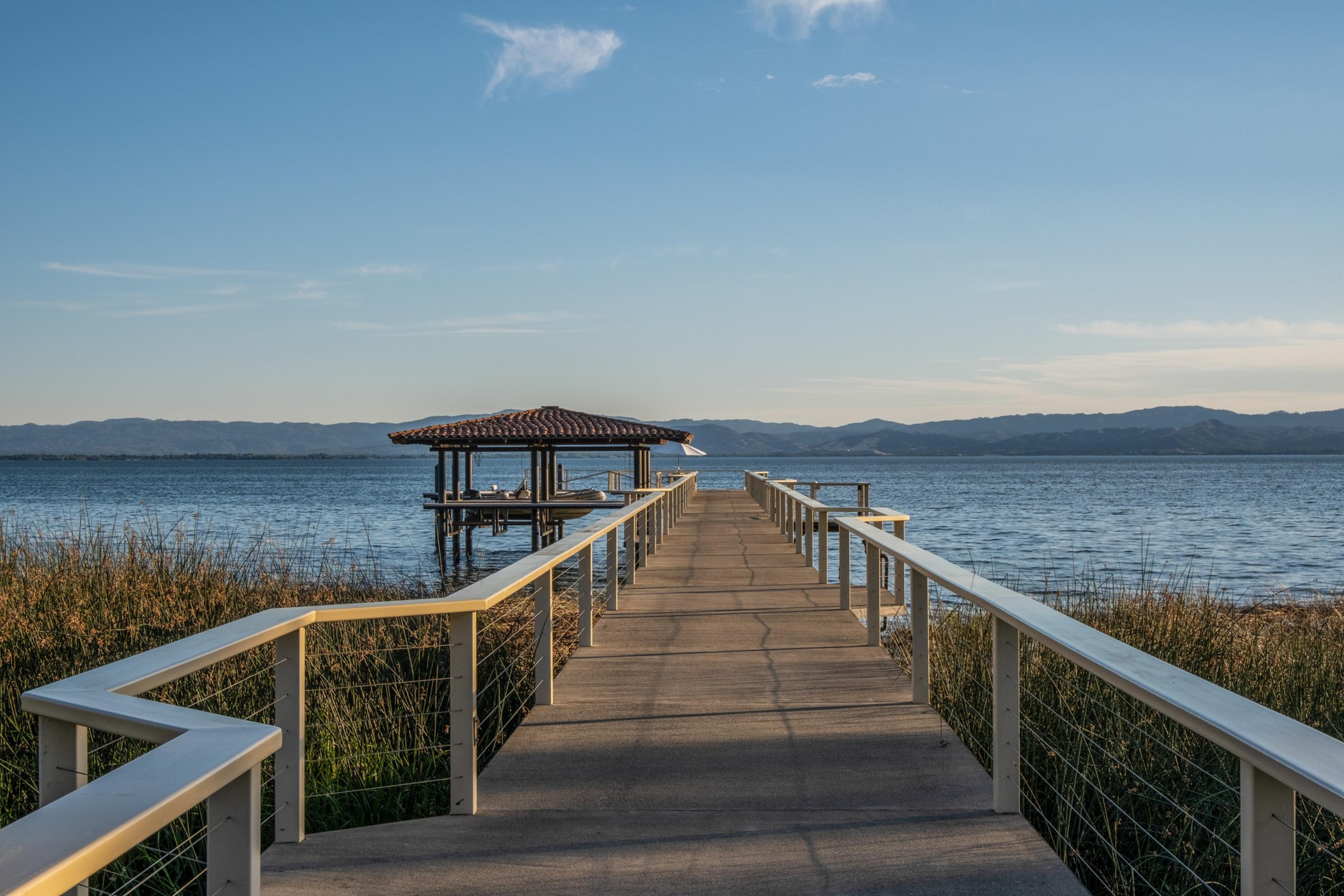 A wooden pier with railings extends over calm water toward a covered dock with a tiled roof, surrounded by tall grass and distant hills under a clear sky.