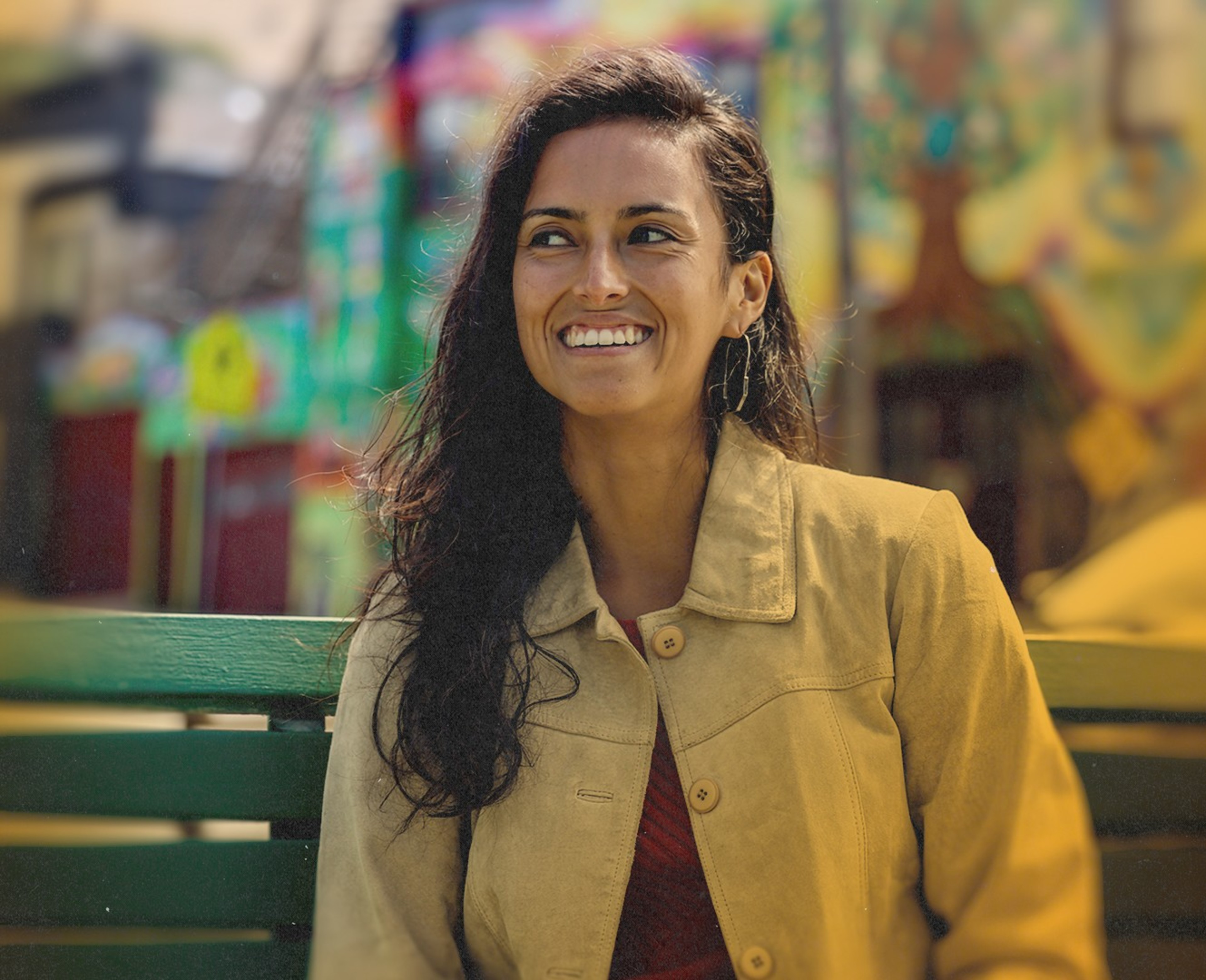 A woman with long dark hair is smiling brightly while sitting on a green bench. She wears a tan jacket, and behind her is a colorful, blurred background.