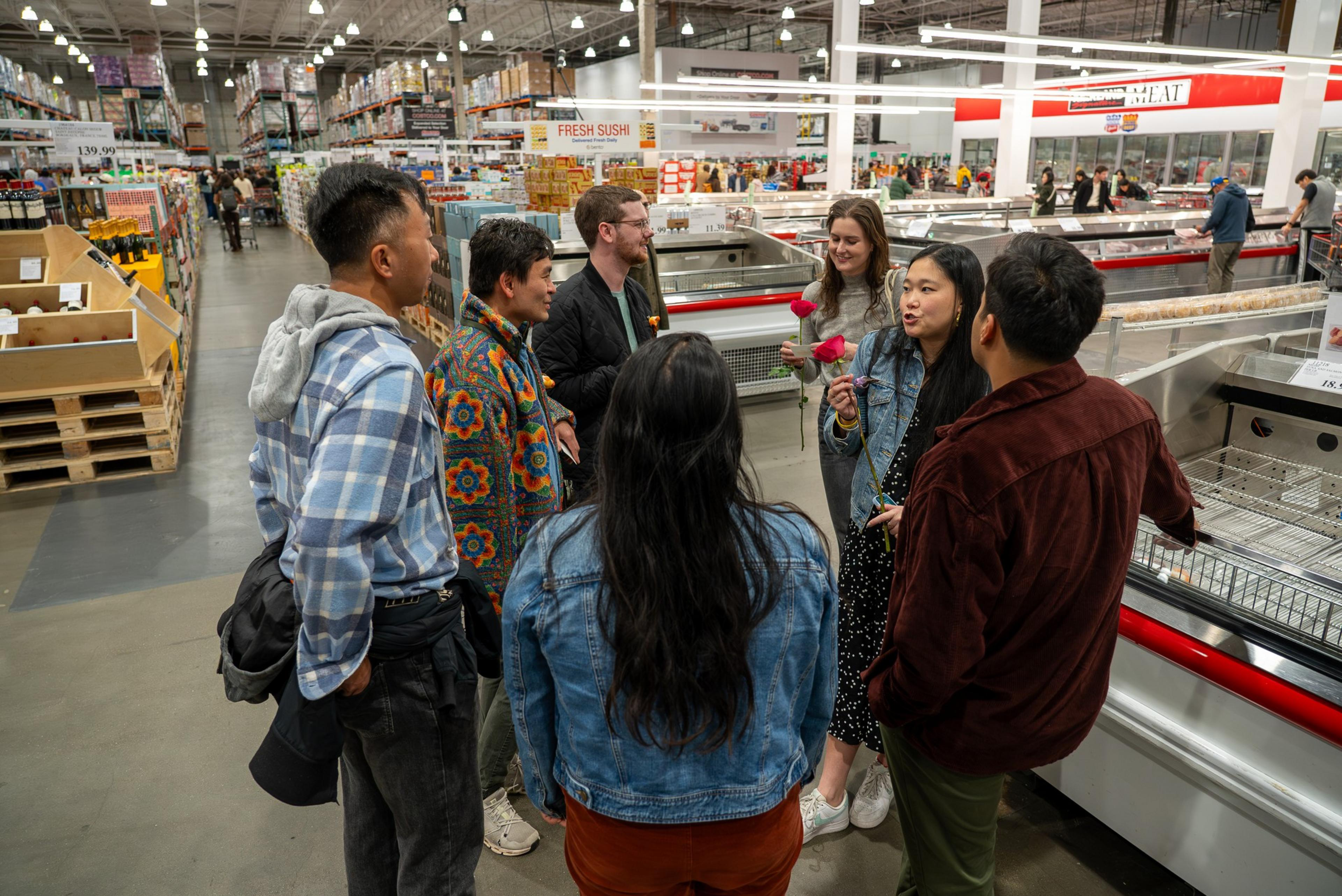 A group of seven people stand and chat near the meat and fresh sushi sections inside a large warehouse store.