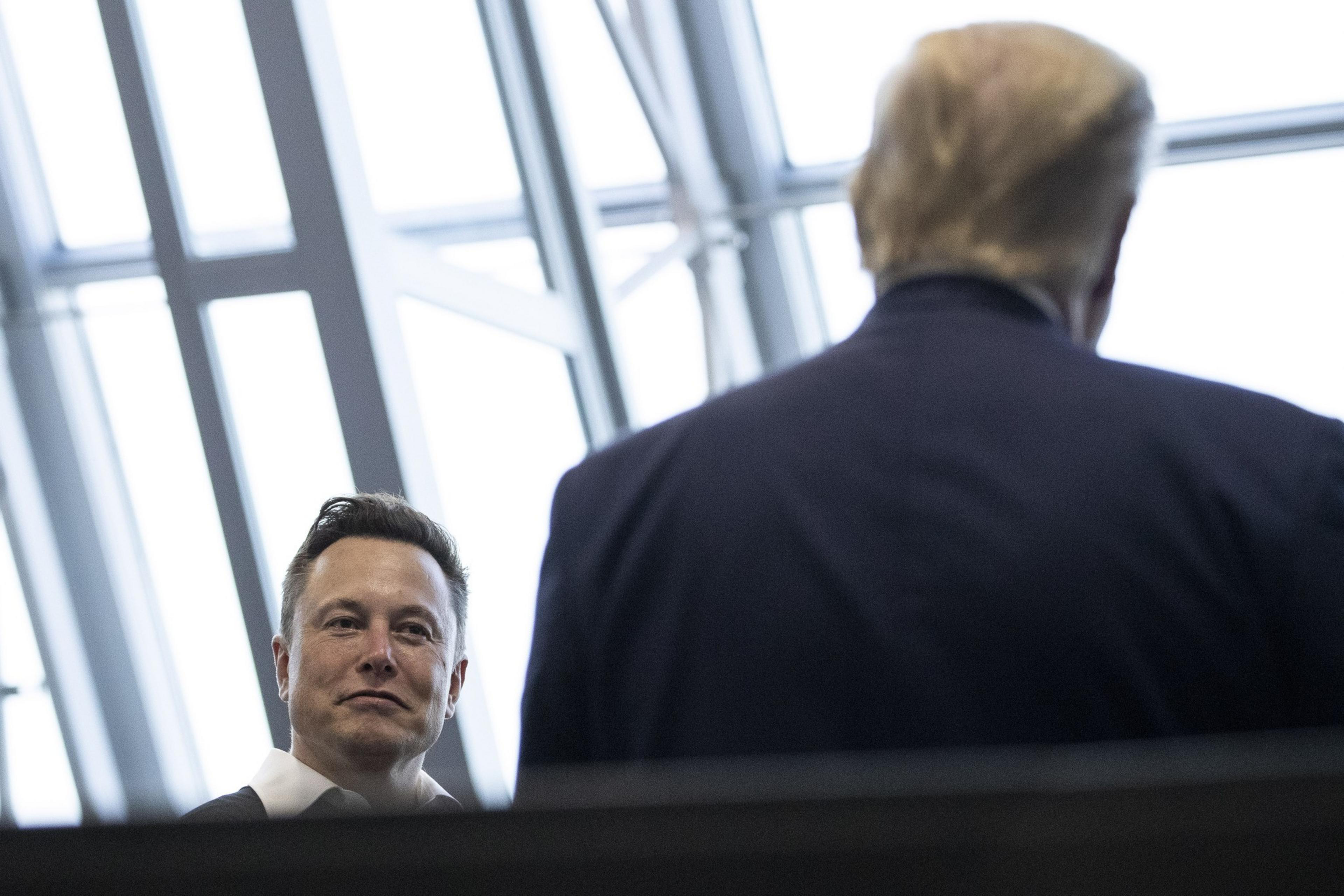 A man with short hair, wearing a white shirt, looks up at another man in a dark suit standing with his back to the camera, in a room with large windows.