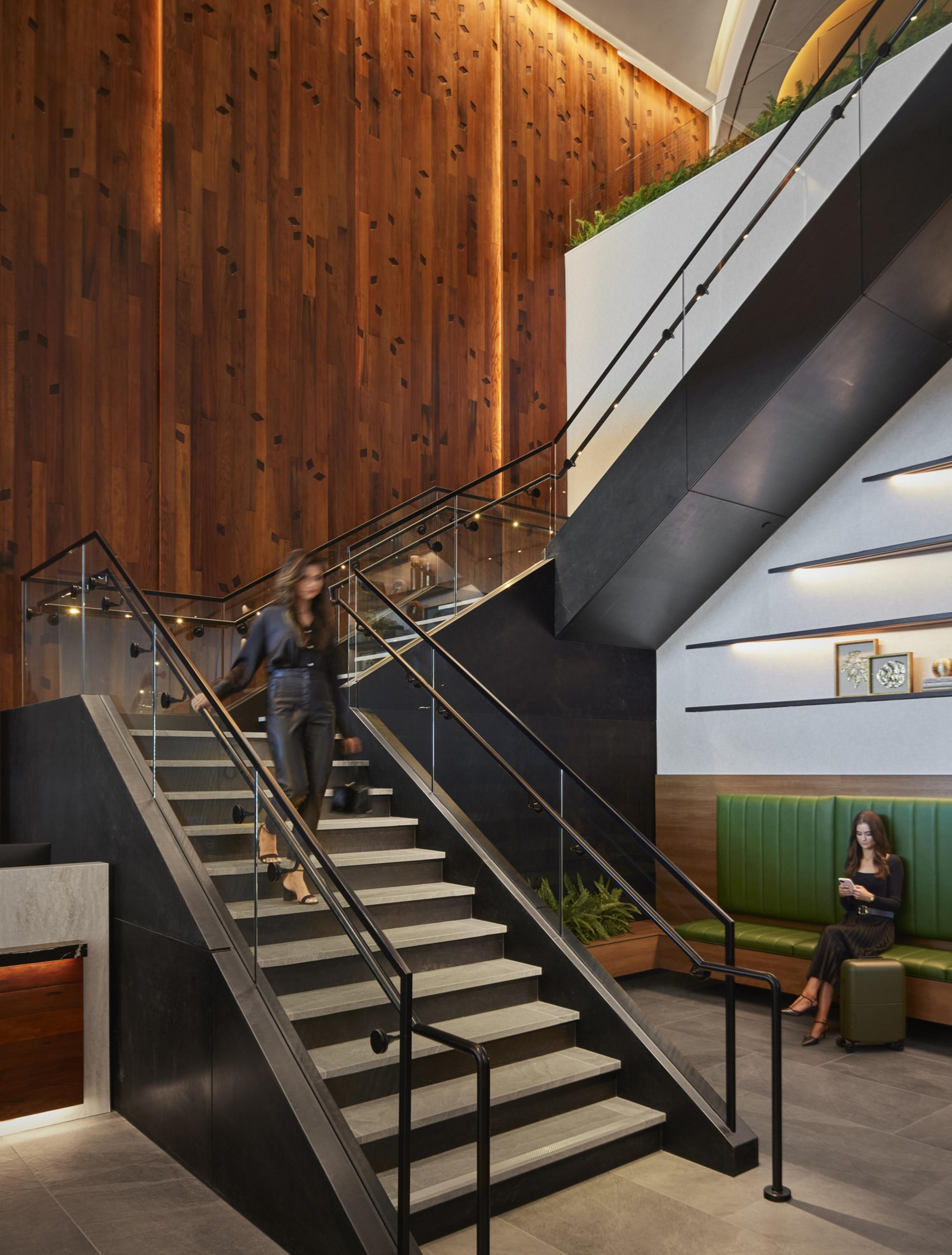 A modern lobby features a person descending a central staircase, a person seated on a green bench looking at their phone, and a warm wooden wall backdrop with plants.