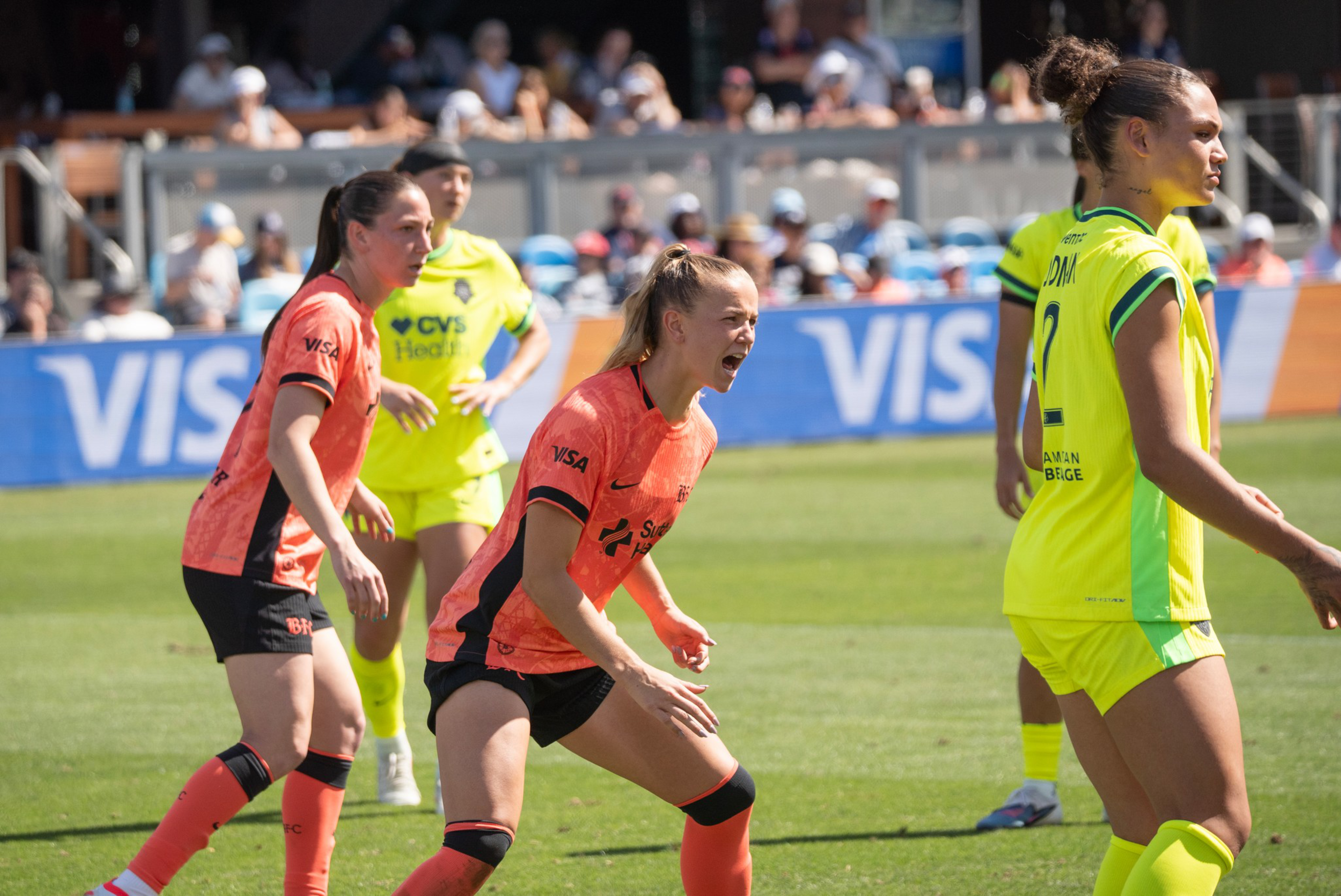 A female soccer player in an orange jersey yells passionately while two teammates and two opponents in yellow watch on a sunny field.