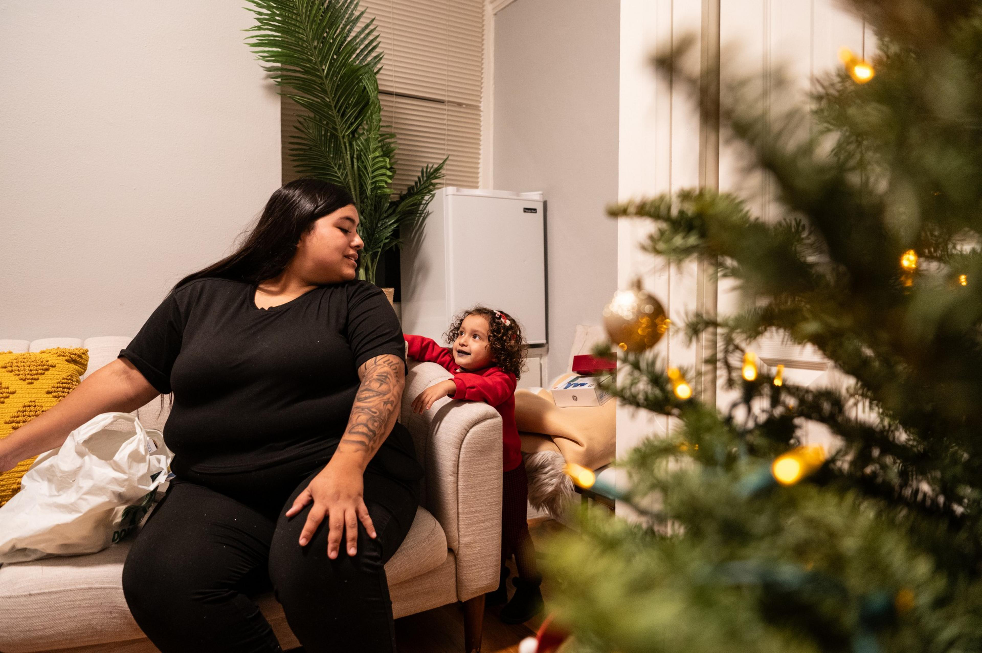 A woman in black sits on a sofa beside a child smiling at her. A Christmas tree with lights and ornaments is in the foreground, creating a festive atmosphere.