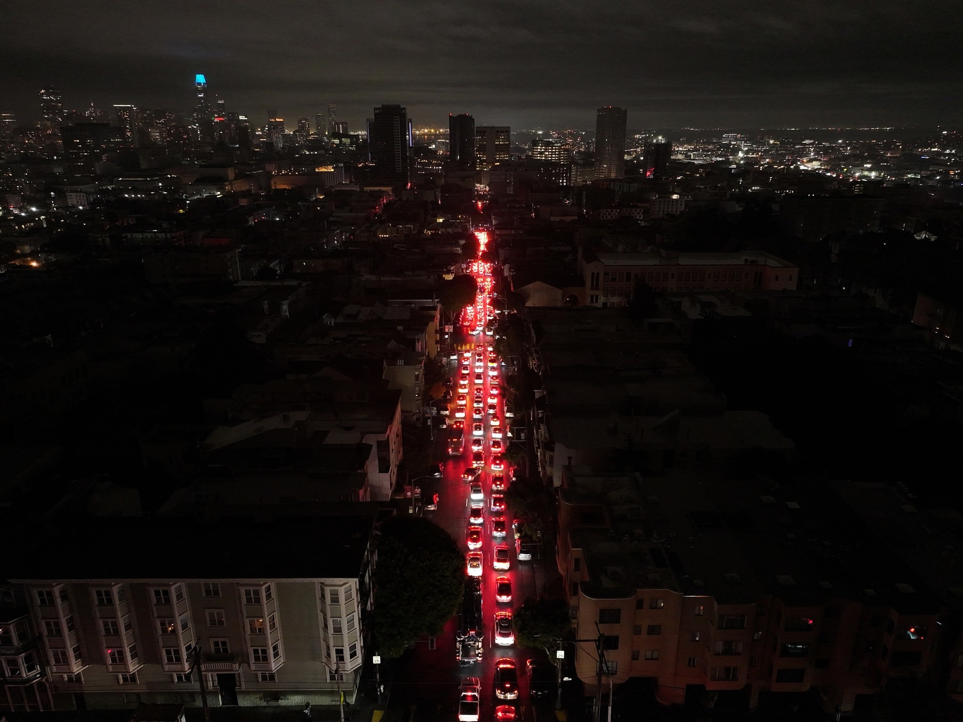 A long line of cars with red brake lights forms a nighttime traffic jam on a city street surrounded by dark buildings and a distant city skyline.