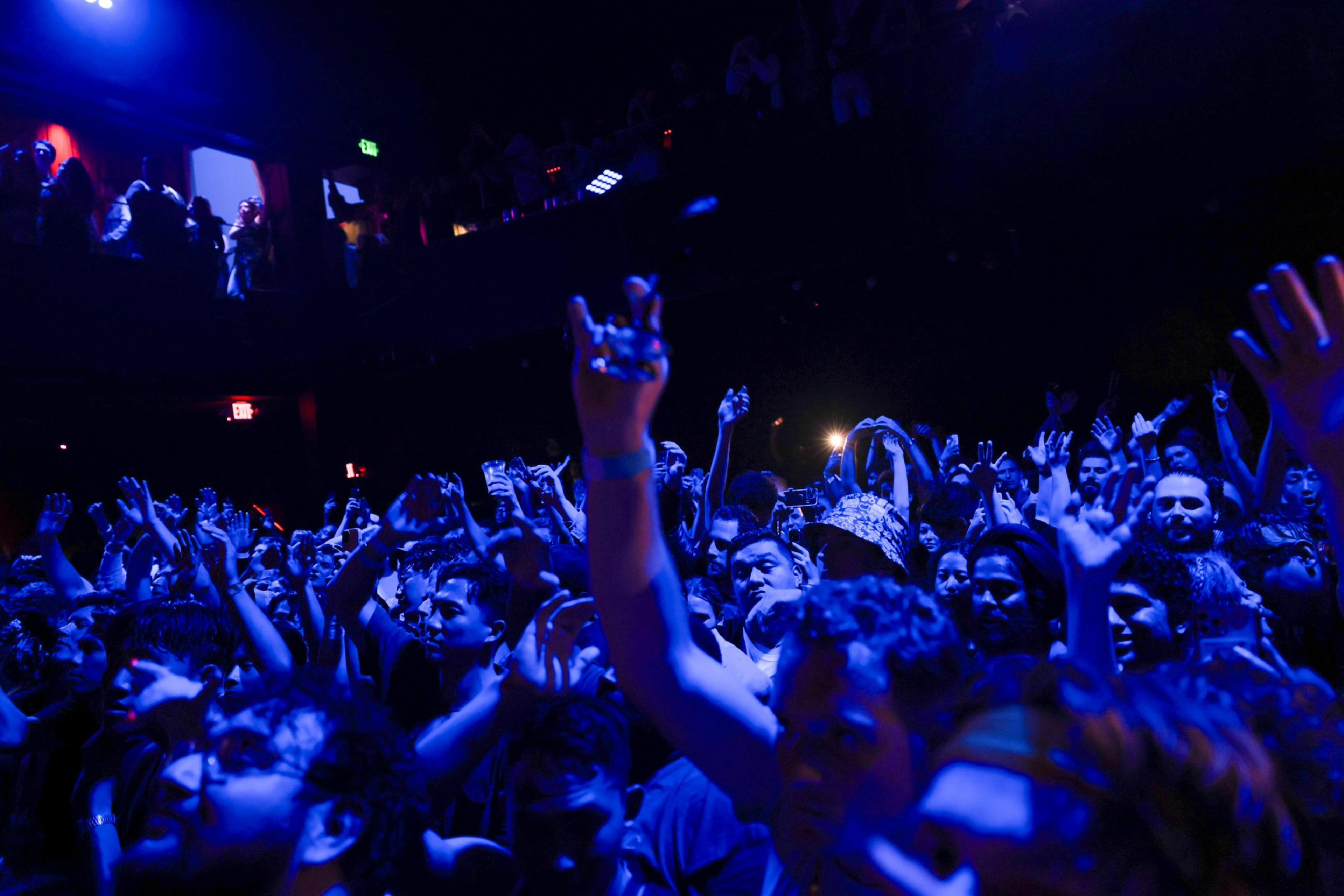 A crowd of people at a concert raise their hands, illuminated by blue stage lights, with some making hand gestures and faces showing excitement.