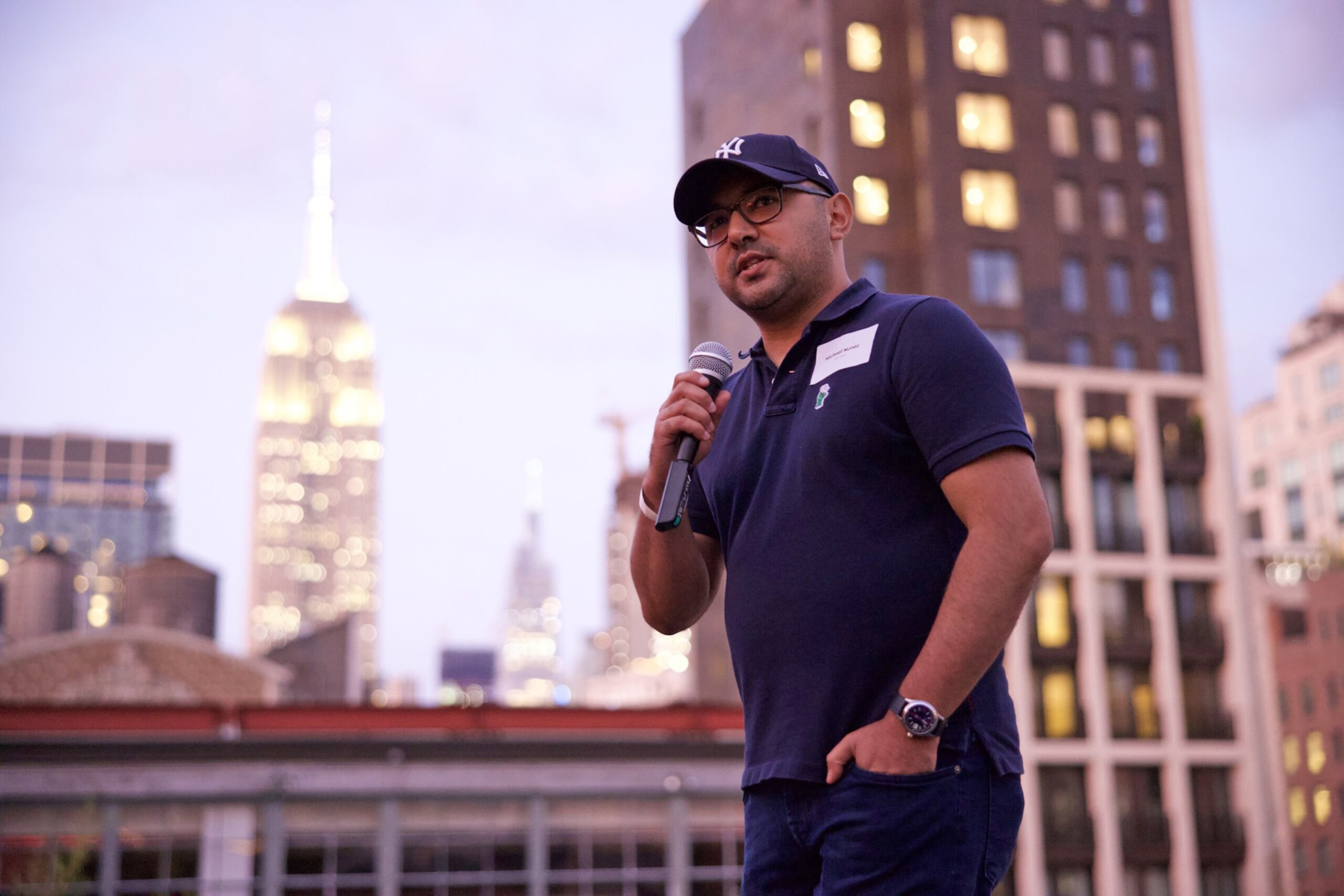 A man wearing a navy blue shirt and cap holds a microphone, speaking outdoors at dusk with city buildings and the Empire State Building lit in the background.