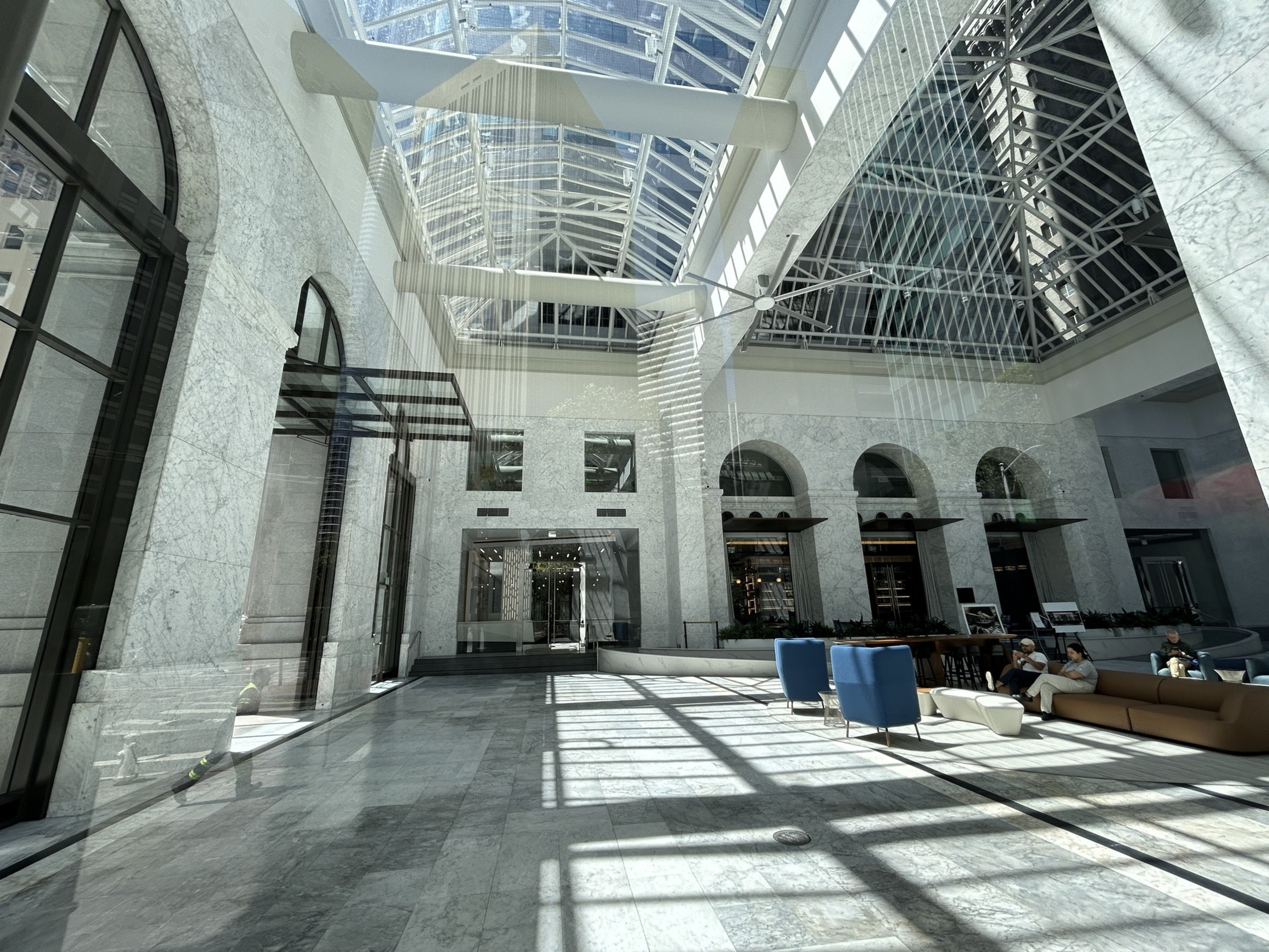 A spacious, modern atrium with a glass ceiling letting in natural light, marble walls, and a seating area with blue and brown furniture.