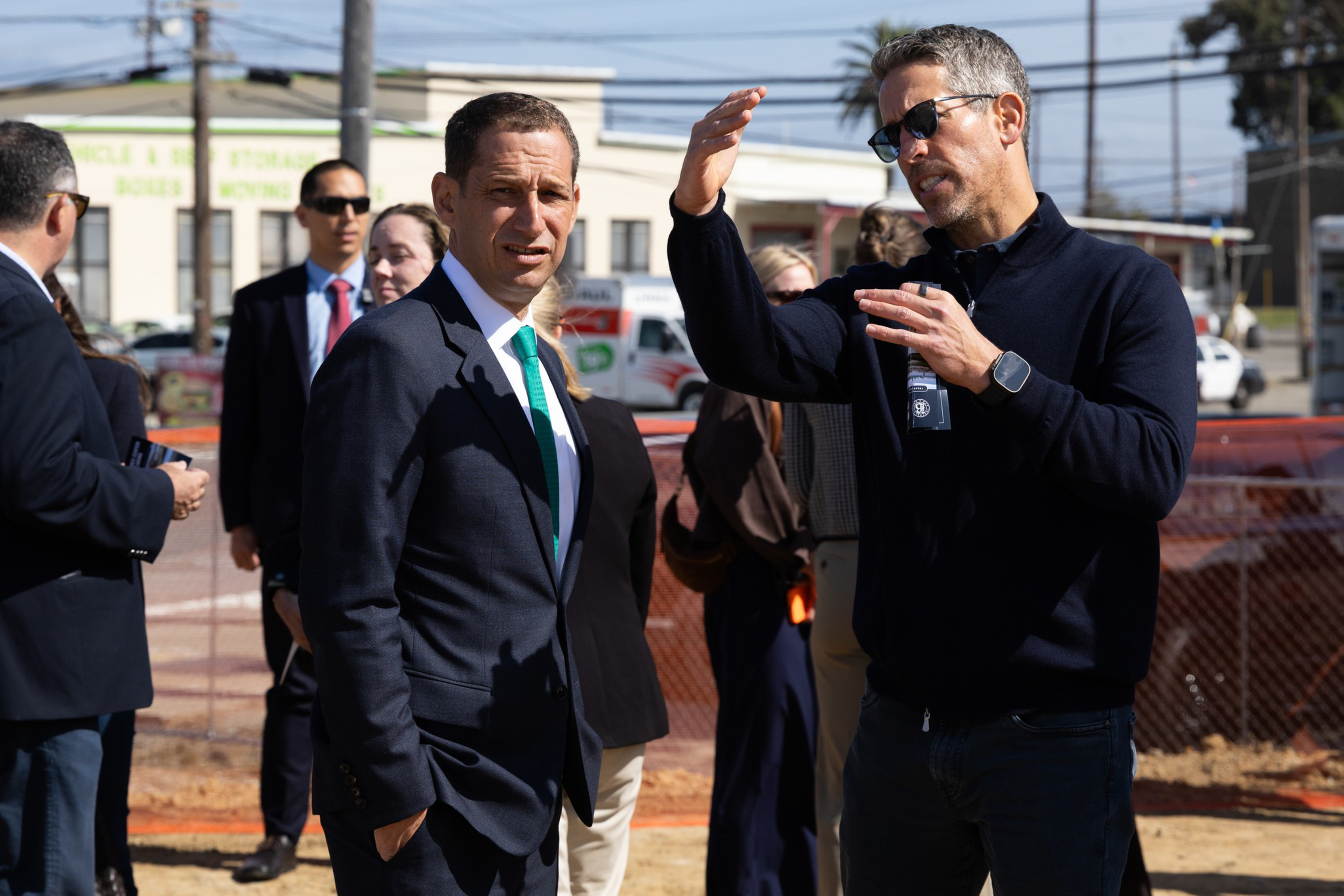 A group of people, including a man in a suit and another man wearing sunglasses and a dark sweater, stand outdoors discussing near a construction site.