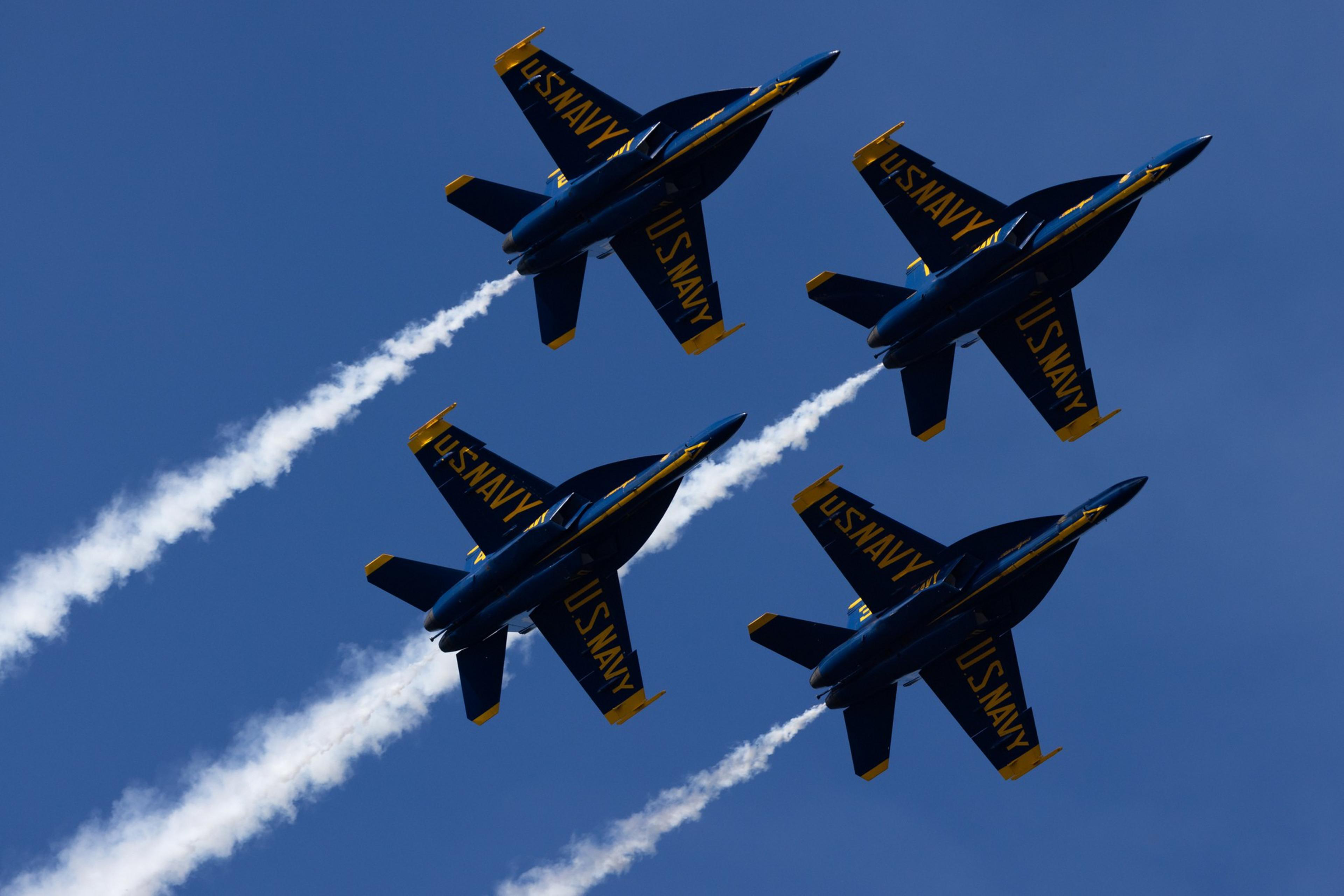 Four fighter jets are flying in a close formation against a clear blue sky, leaving white vapor trails behind them. "U.S. Navy" is visible on the aircrafts.