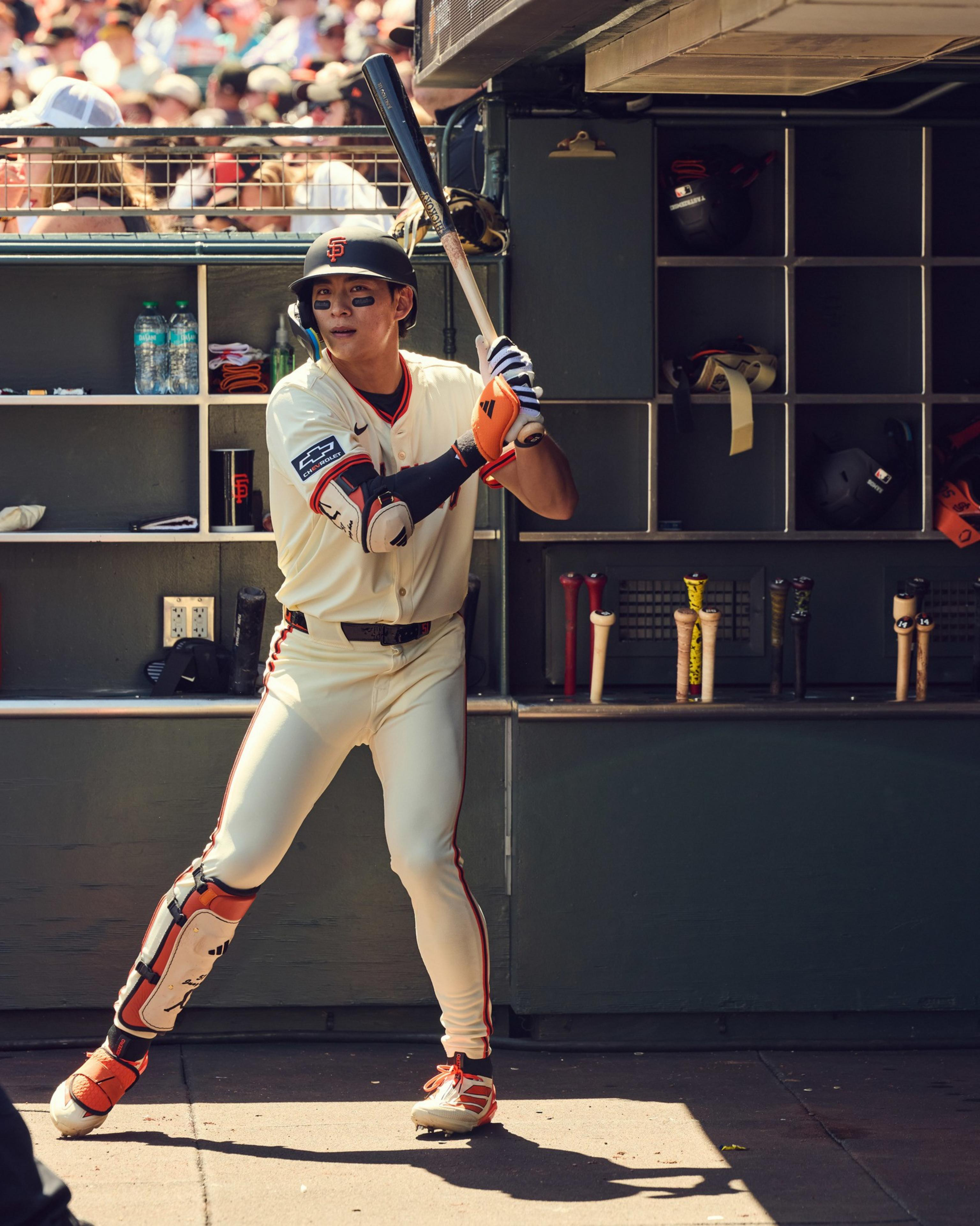 A baseball player in a white uniform and helmet stands ready to bat near the dugout. He wears protective gear and is surrounded by equipment and a crowd in the background.