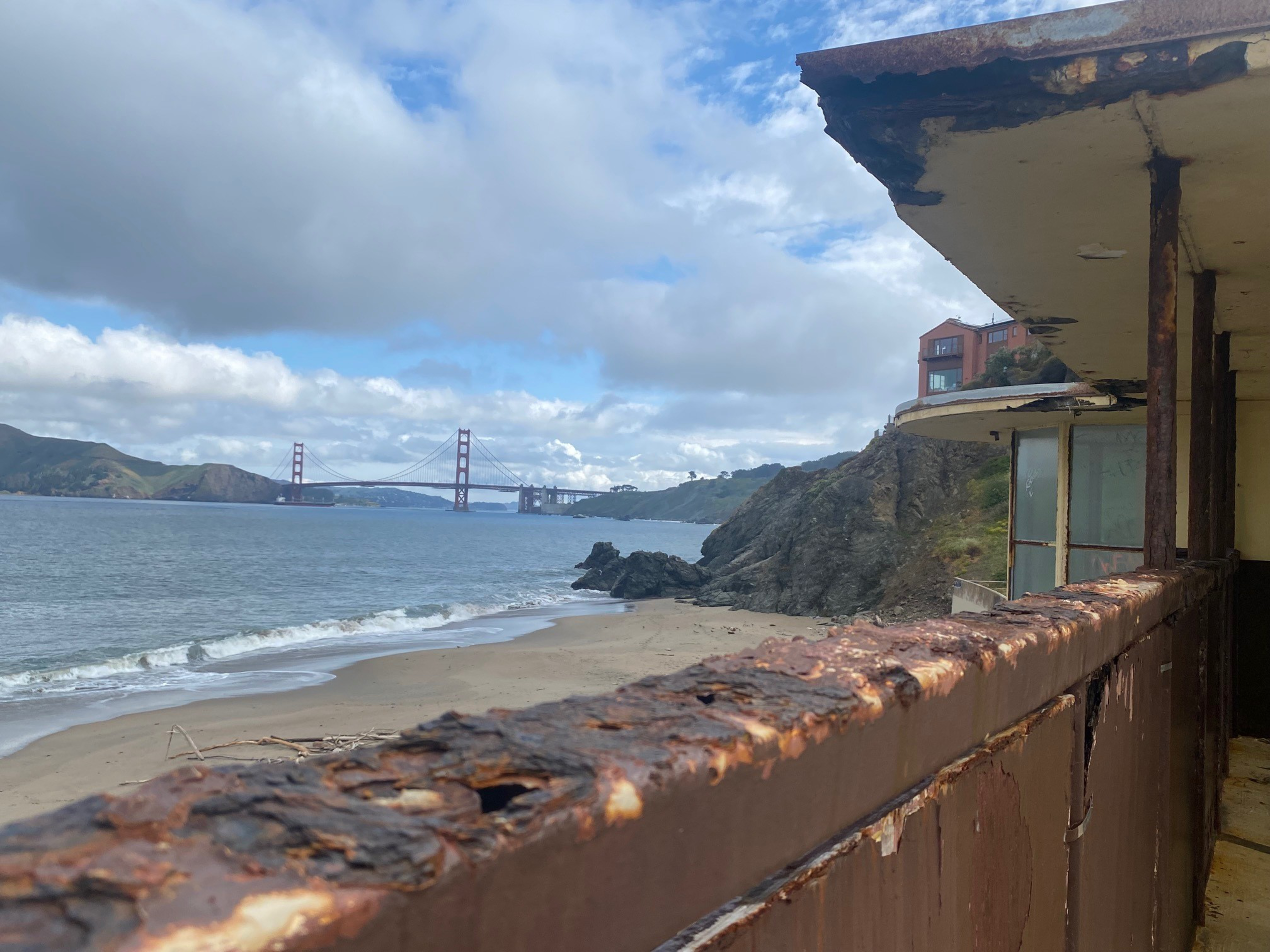 A rusty railing and rundown building overlook a sandy beach and rocky coastline, with the Golden Gate Bridge visible across the water under a cloudy sky.