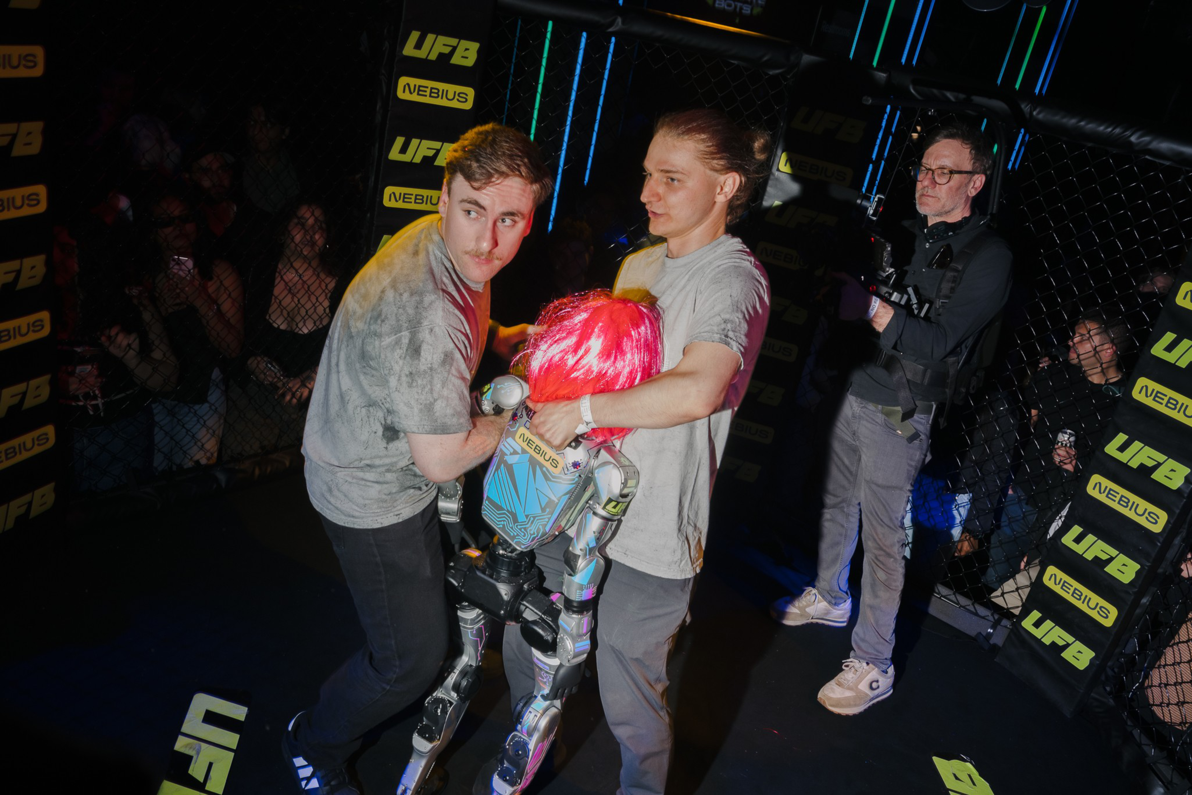 Two men are holding a small humanoid robot with bright pink hair inside a fighting cage, while a cameraman films nearby.