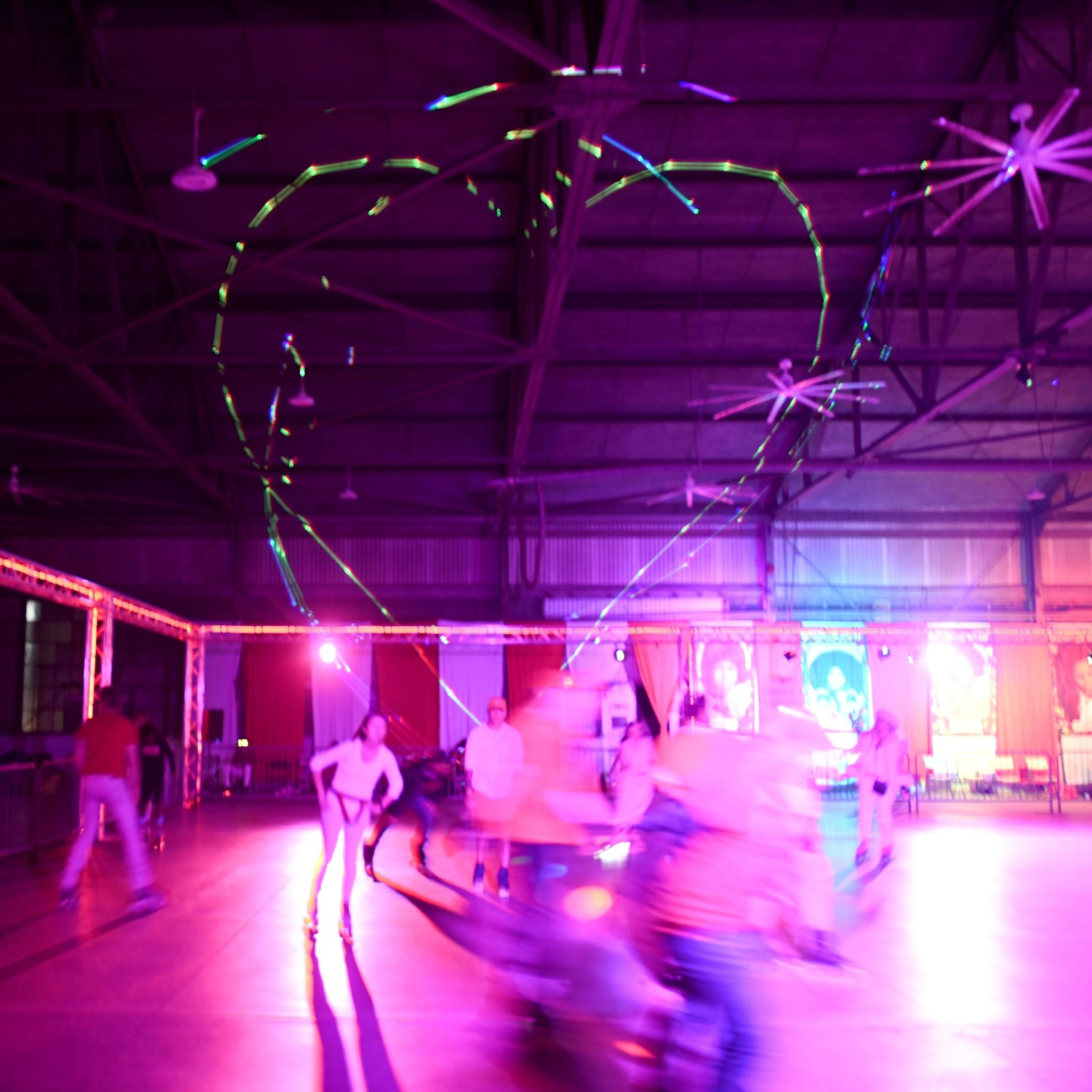 A neon-lit indoor roller-skating rink with blurred motion skaters creating colorful light trails, under a high ceiling with large overhead fans.