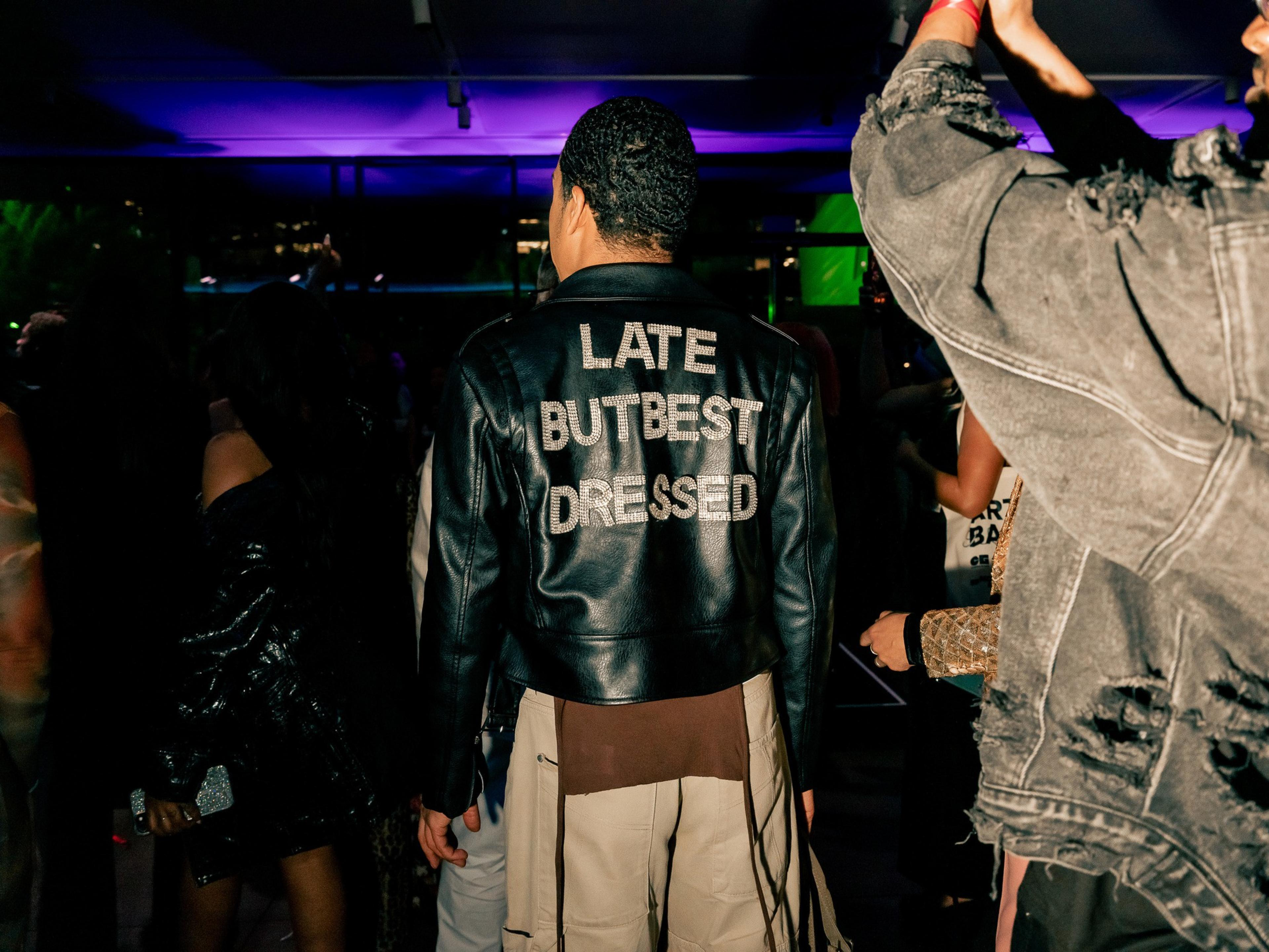 A person wears a black leather jacket with &quot;LATE BUT BEST DRESSED&quot; written on the back. They stand amidst a crowd under purple lights.