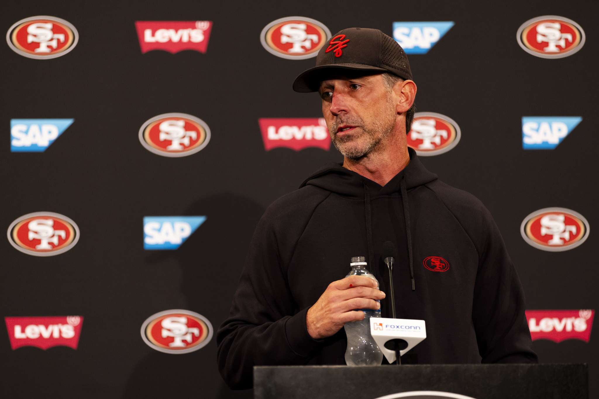 A man in a black hoodie and cap with the San Francisco 49ers logo holds a water bottle while speaking at a podium with team and sponsor logos behind him.