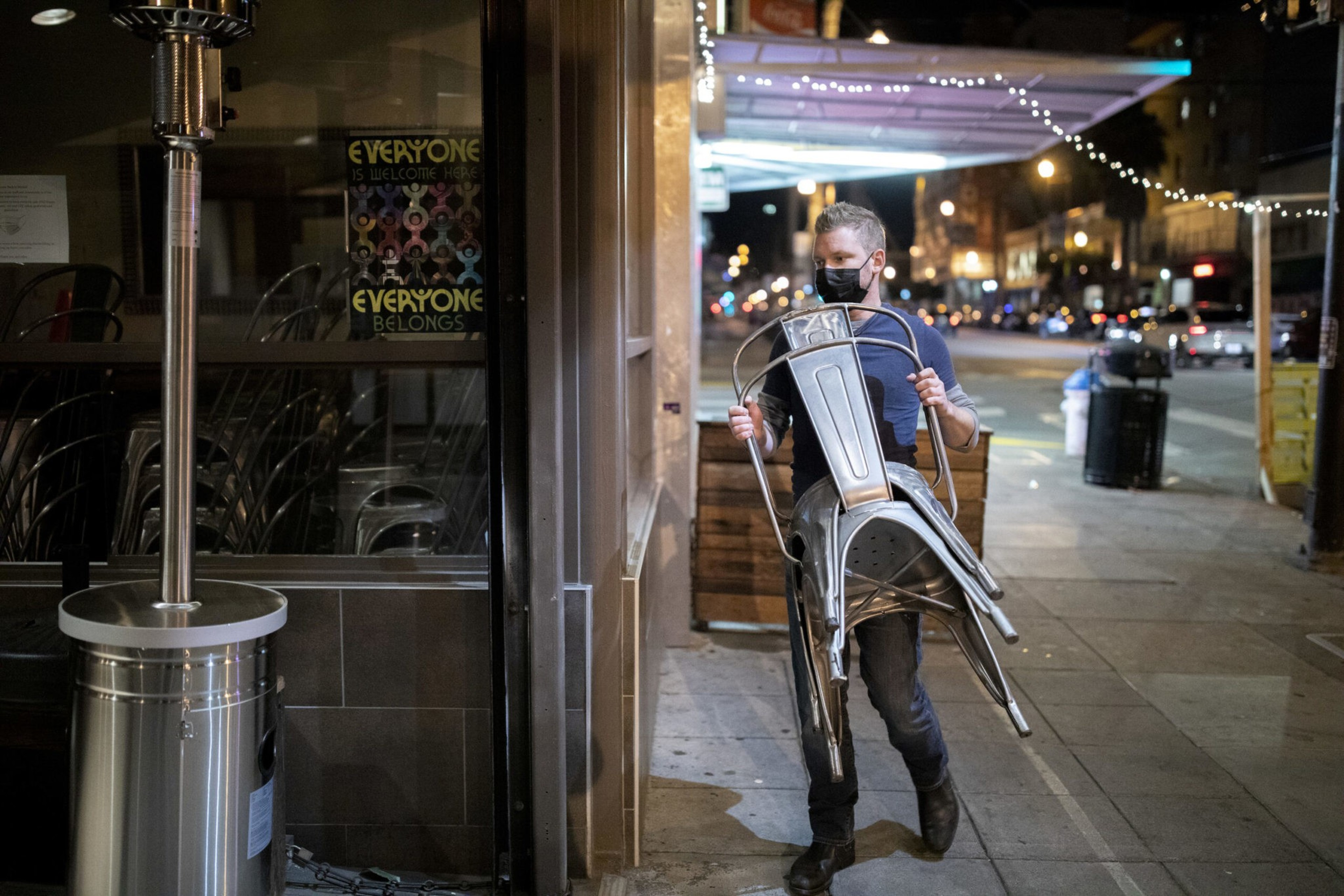 A man wearing a black mask carries three metal chairs on a city sidewalk at night, with streetlights and buildings in the background.
