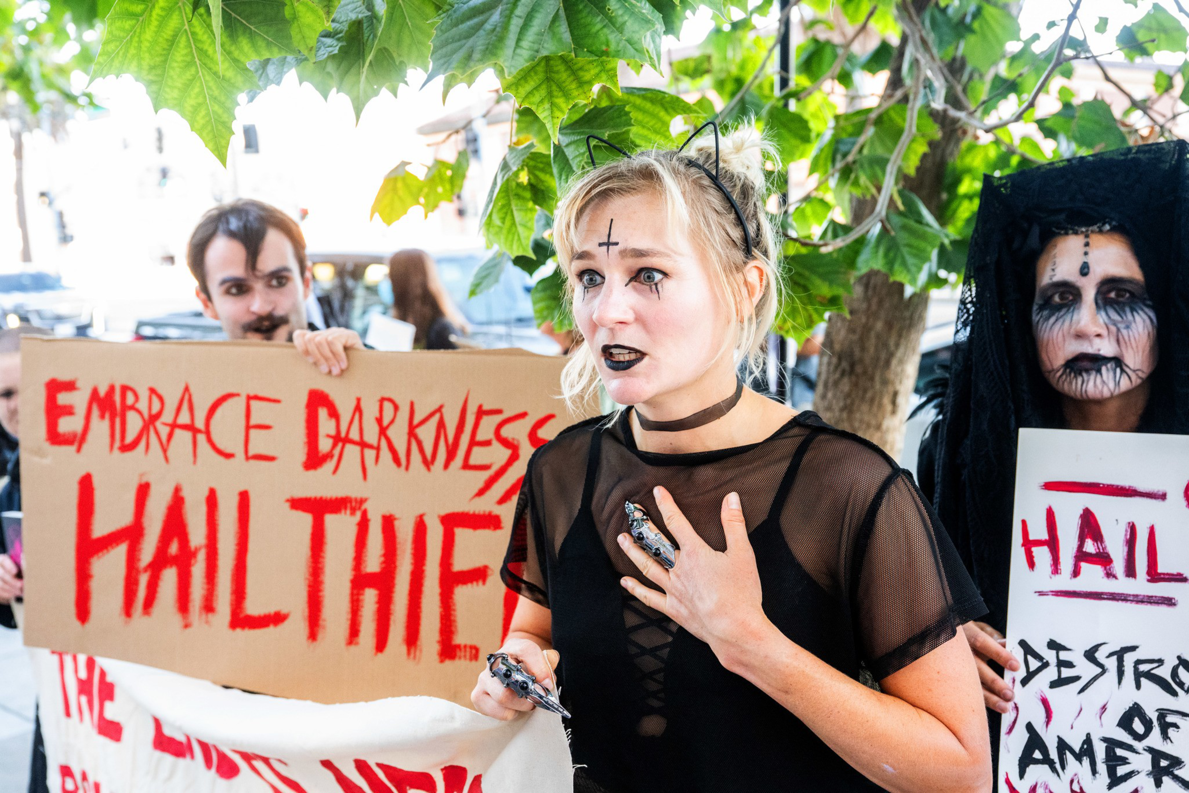 A group of people with gothic makeup and costumes hold signs with red and black text promoting dark themes and rebellion.