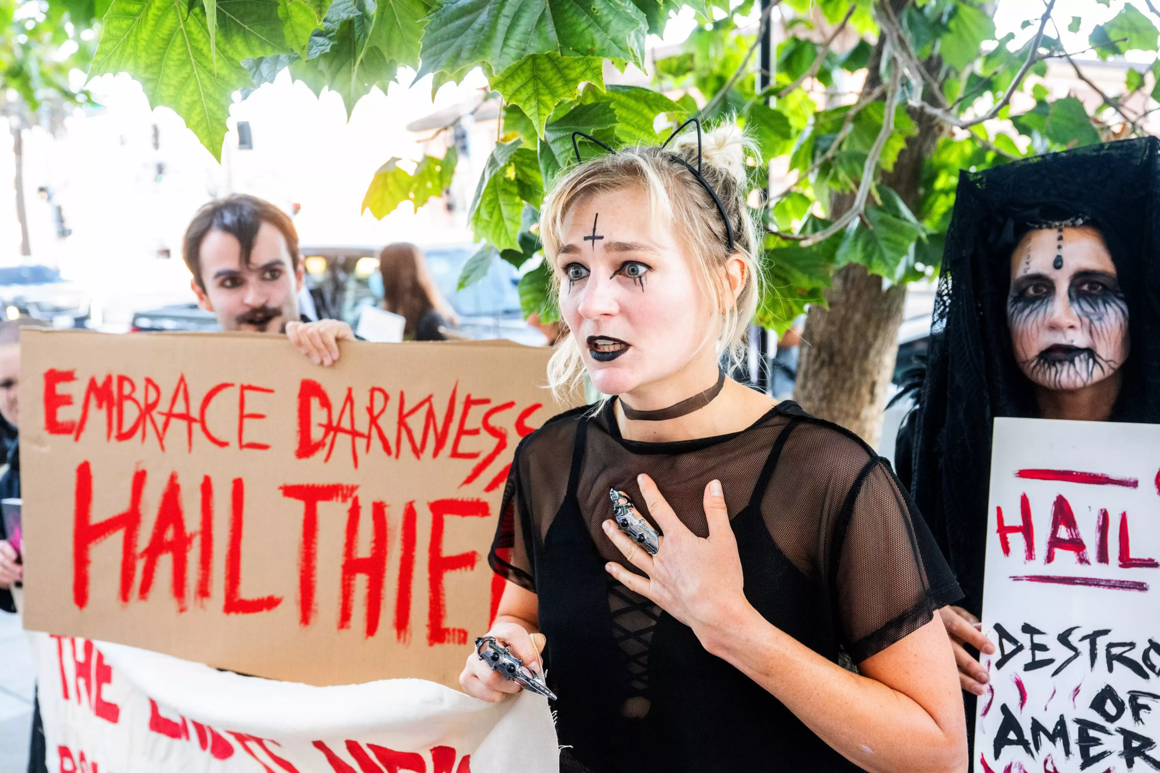 A group of people with gothic makeup and costumes hold signs with red and black text promoting dark themes and rebellion.