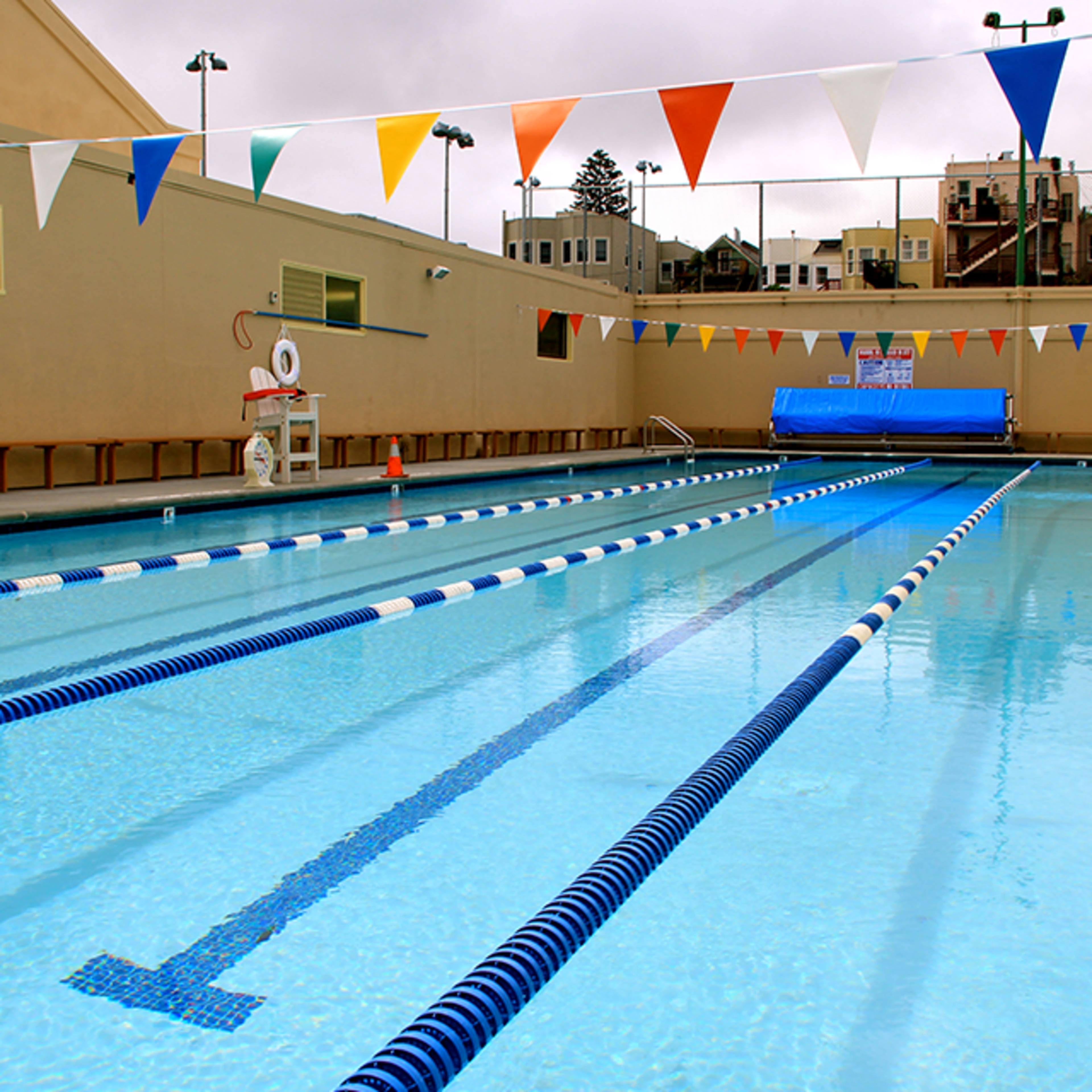 An empty outdoor swimming pool with lane dividers, colorful triangular flags overhead, a lifeguard chair, and buildings in the background.