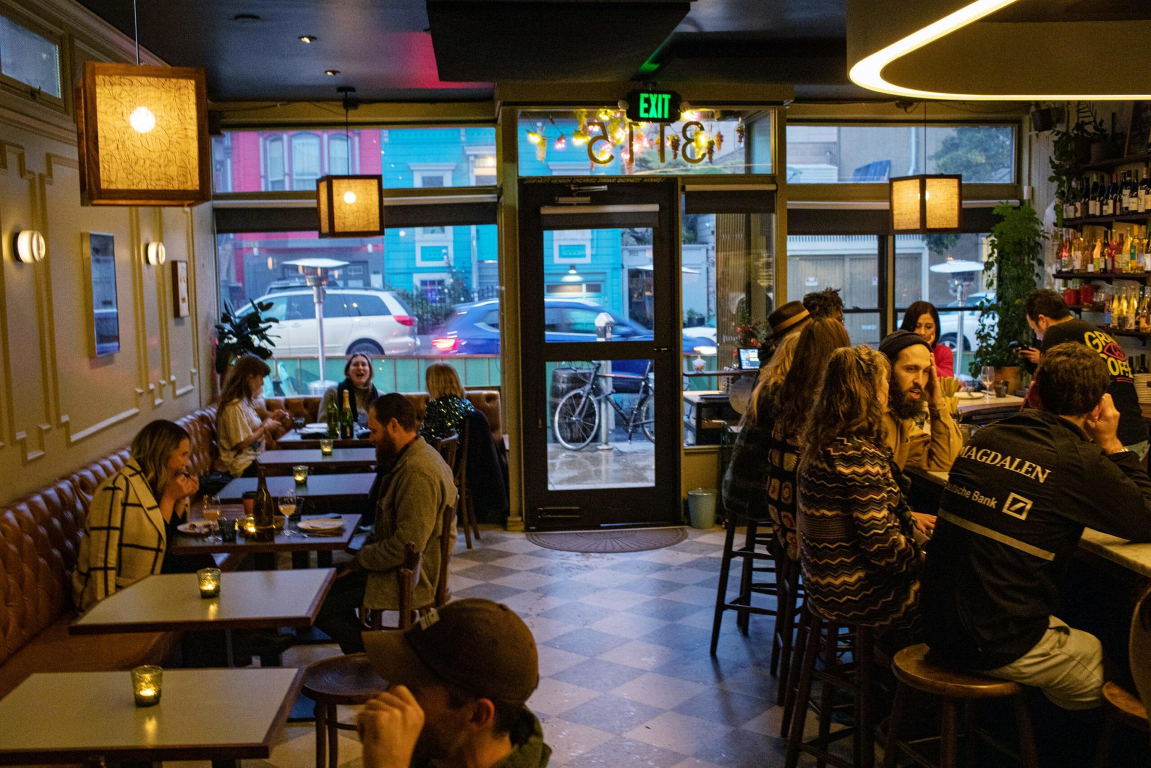 Several people sit at tables and a bar inside a cozy, dimly lit café with large windows showing parked cars and a bike outside.