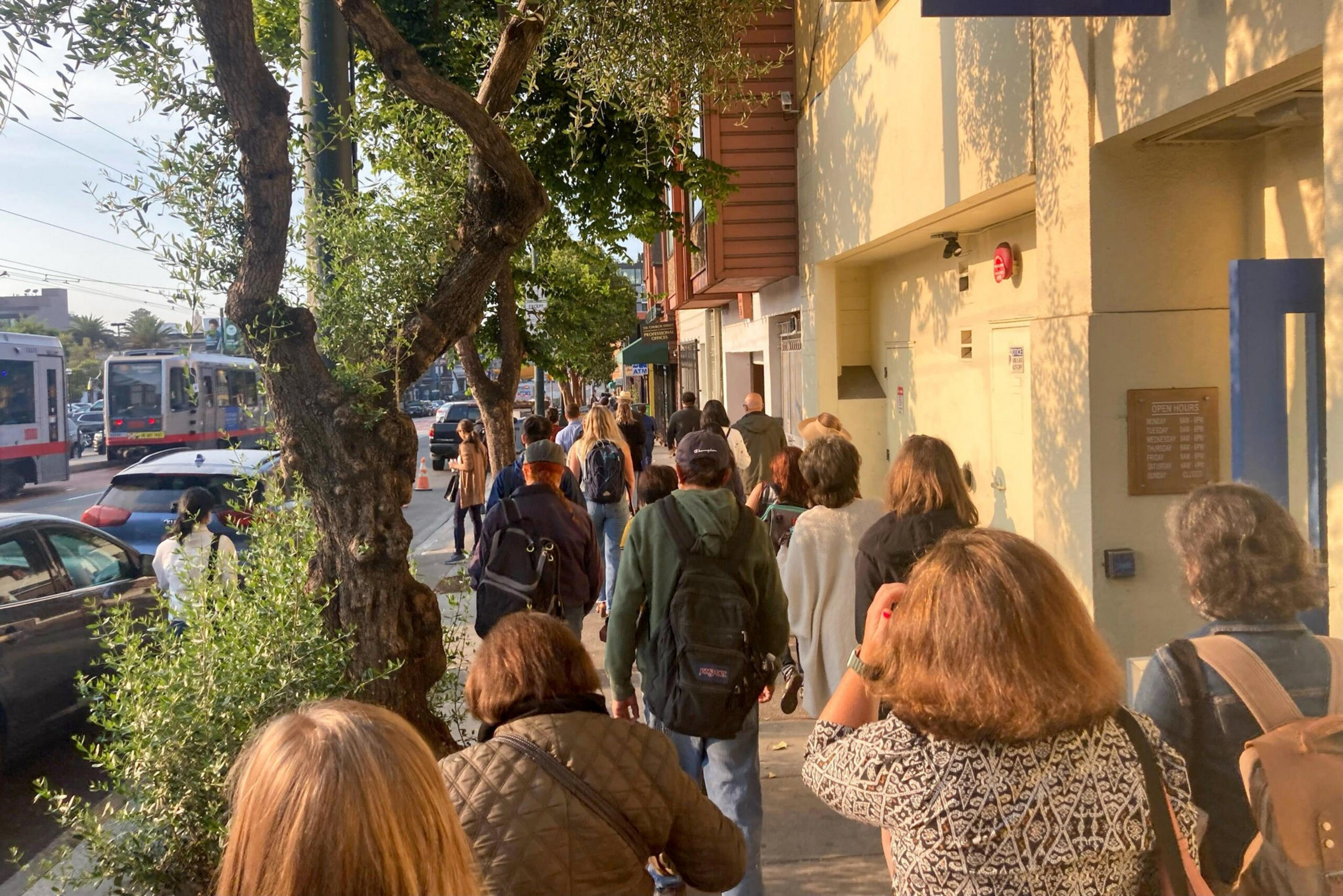 A crowd of people walks along a sunlit city sidewalk lined with trees and buildings, with cars and buses visible on the adjacent street.