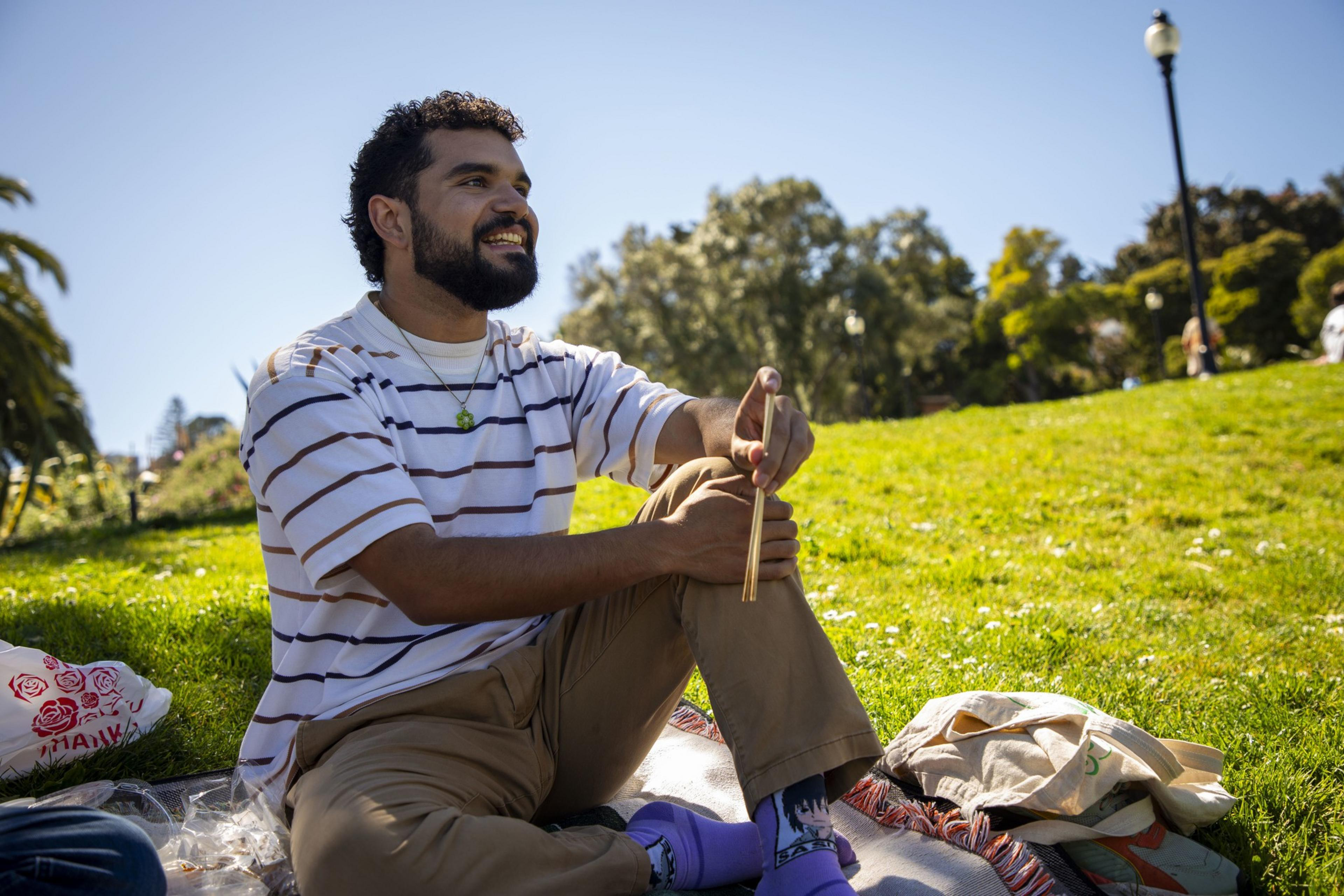 A person sits on a blanket in a sunny park, smiling and holding chopsticks. They wear a striped shirt, brown pants, and purple socks. A lamp post is in the background.