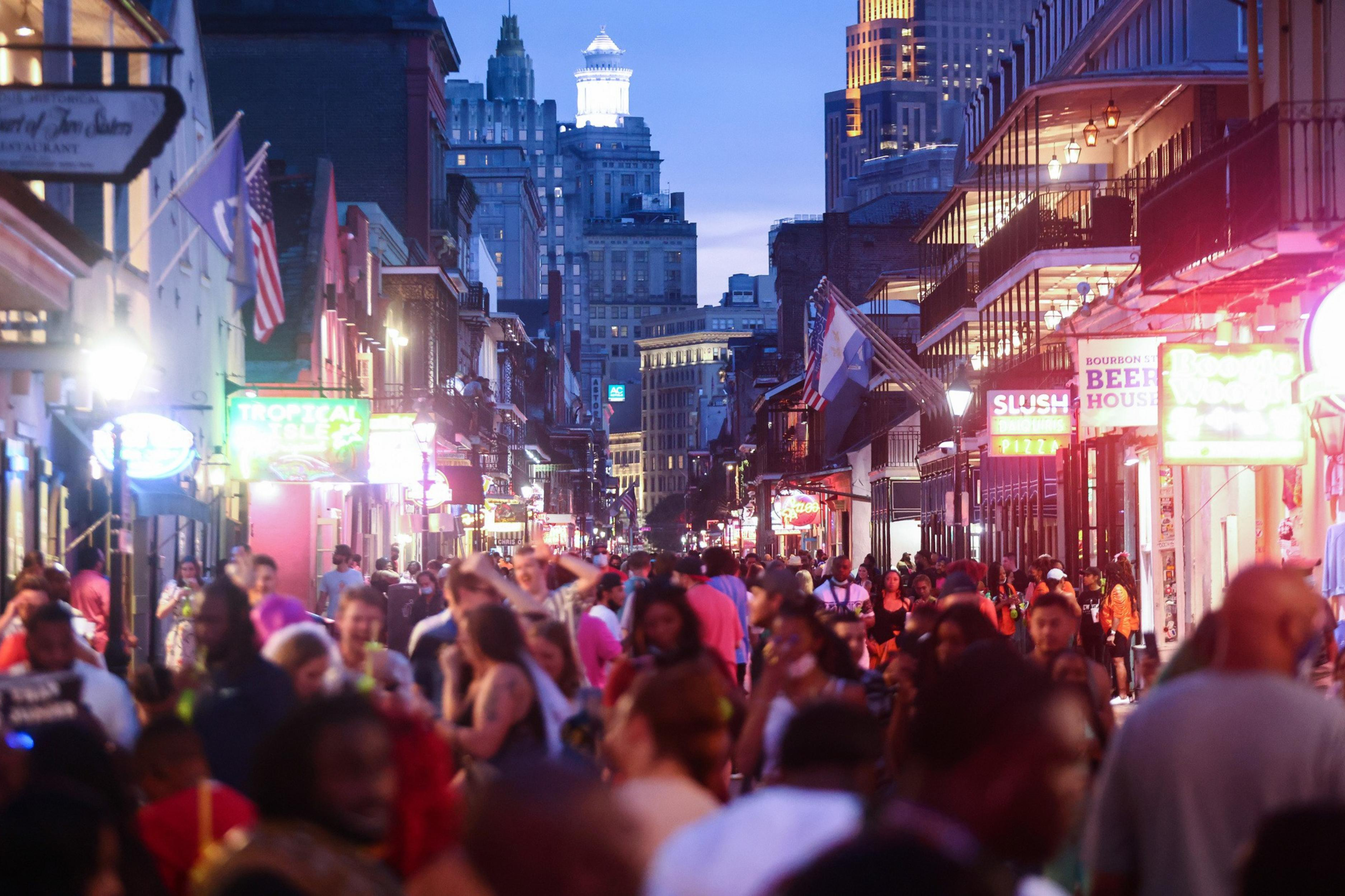 A bustling city street at dusk, crowded with people and lined with neon signs and balconies.