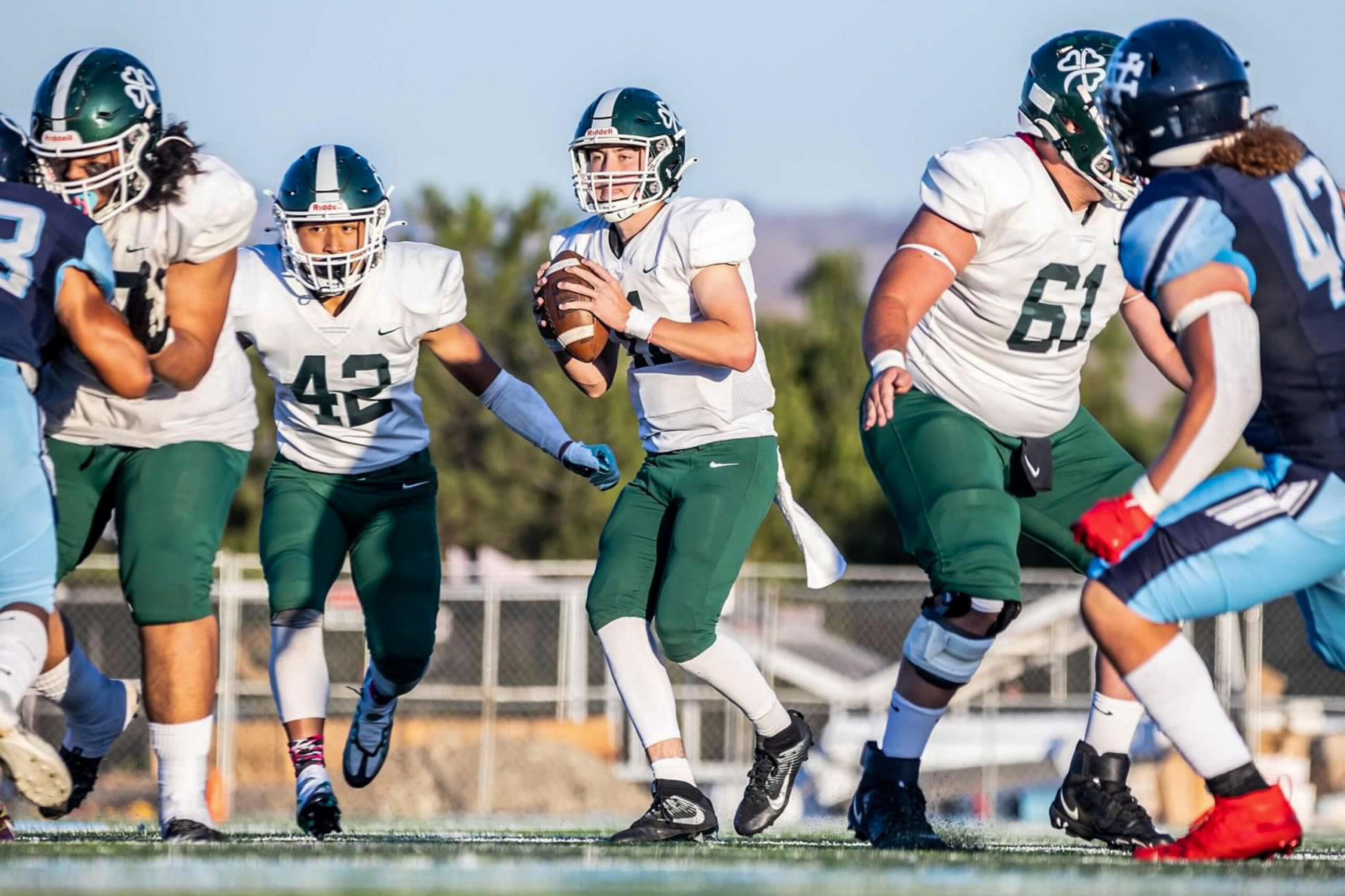 A football quarterback in green and white prepares to throw while teammates block opponents wearing blue and black on a sunny outdoor field.