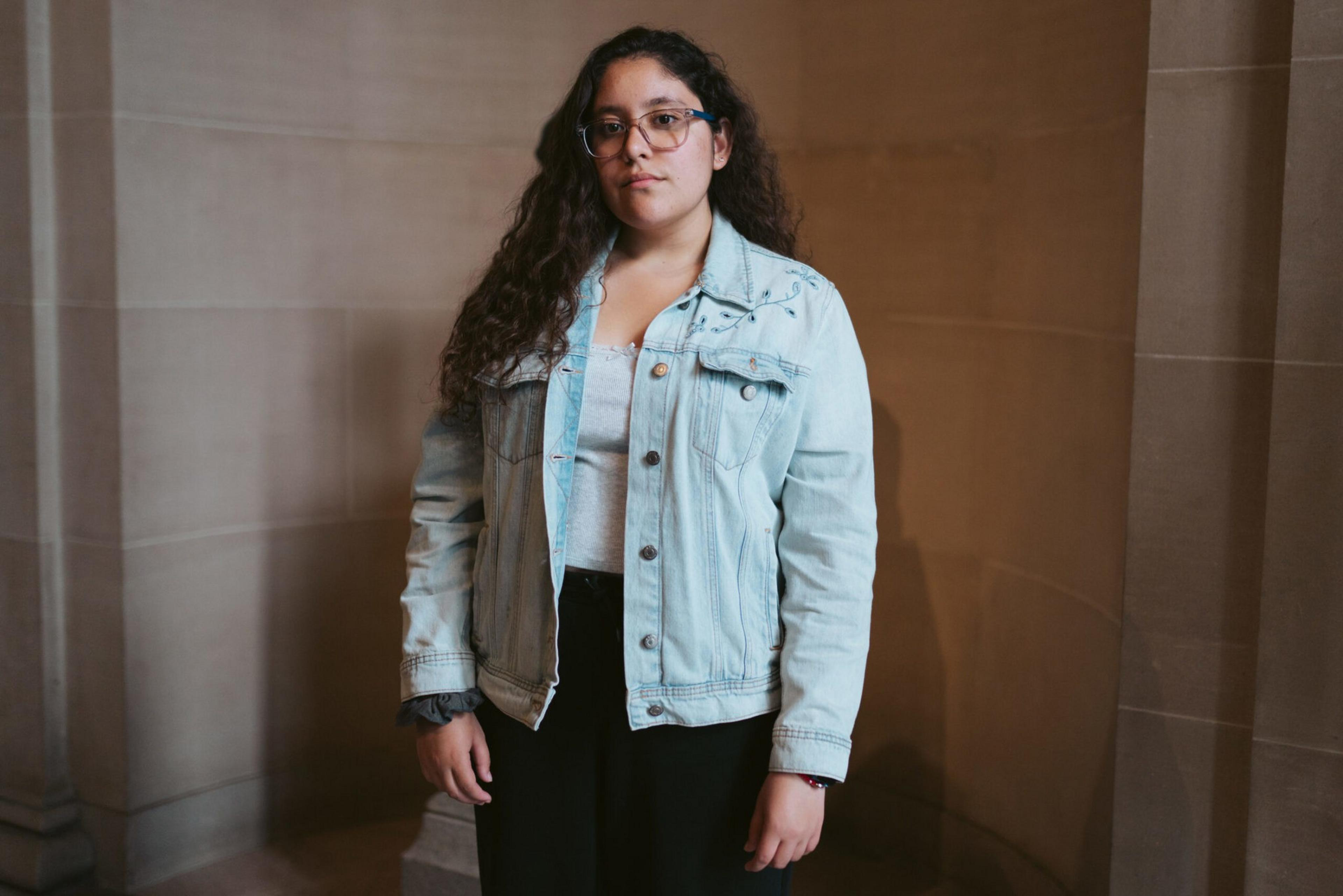 A young woman with long curly hair and glasses wears a light blue denim jacket over a white top, standing against a beige stone wall.