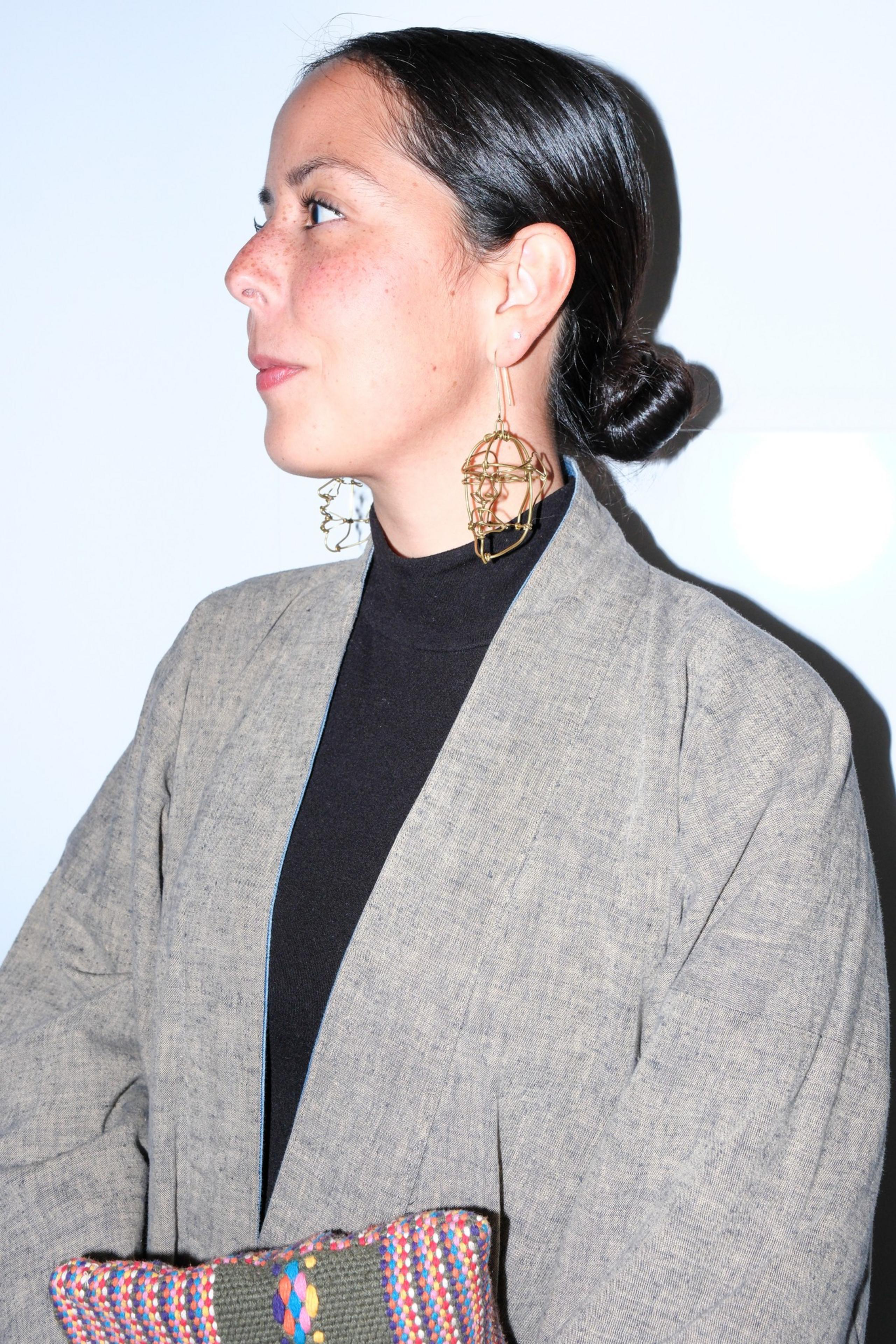 A person in profile with slicked-back hair and bold geometric earrings wears a gray jacket and holds a colorful woven item against a white background.