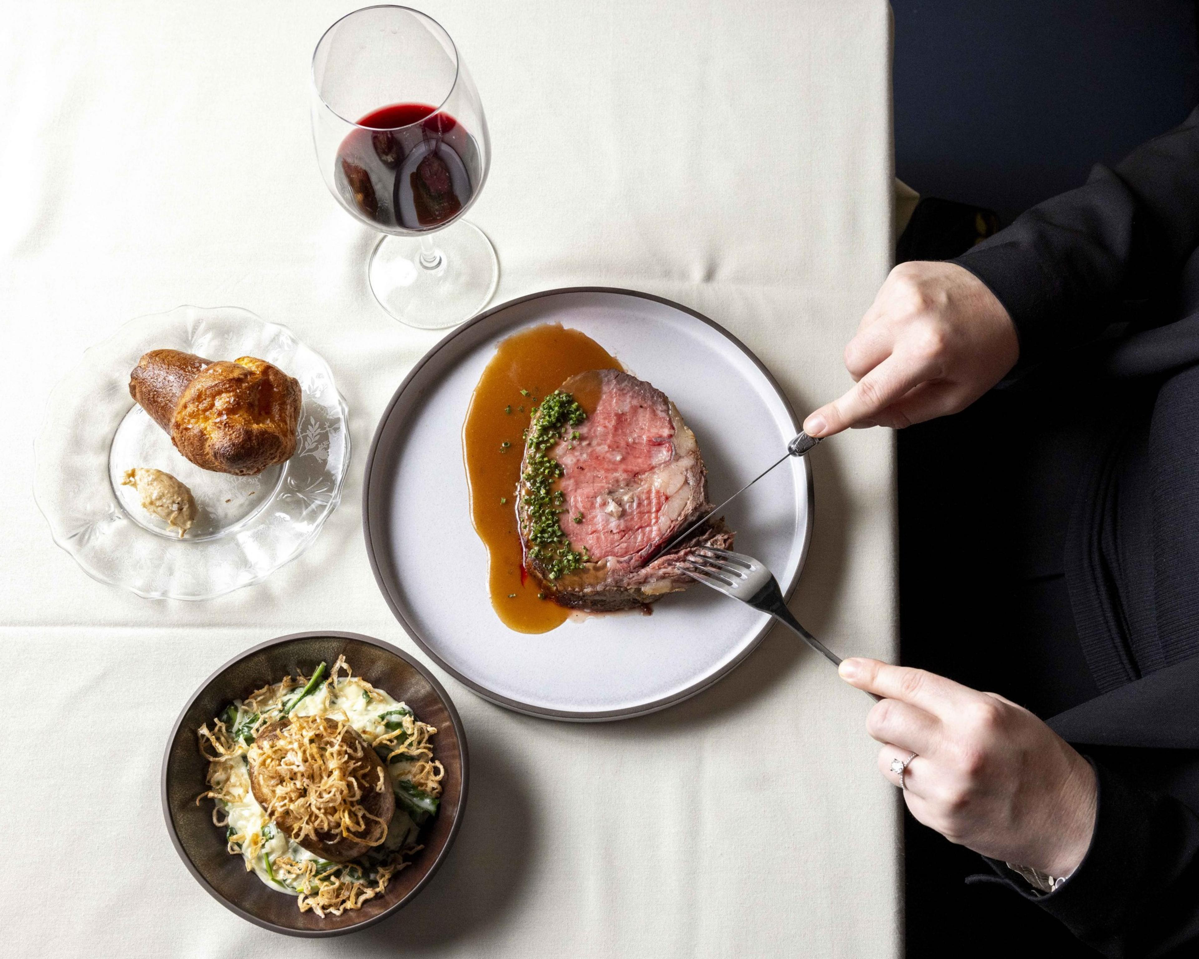 A person is slicing roast beef on a plate with gravy. Nearby are a glass of red wine, a popover with horseradish, and a bowl of creamy green beans with crispy onions.