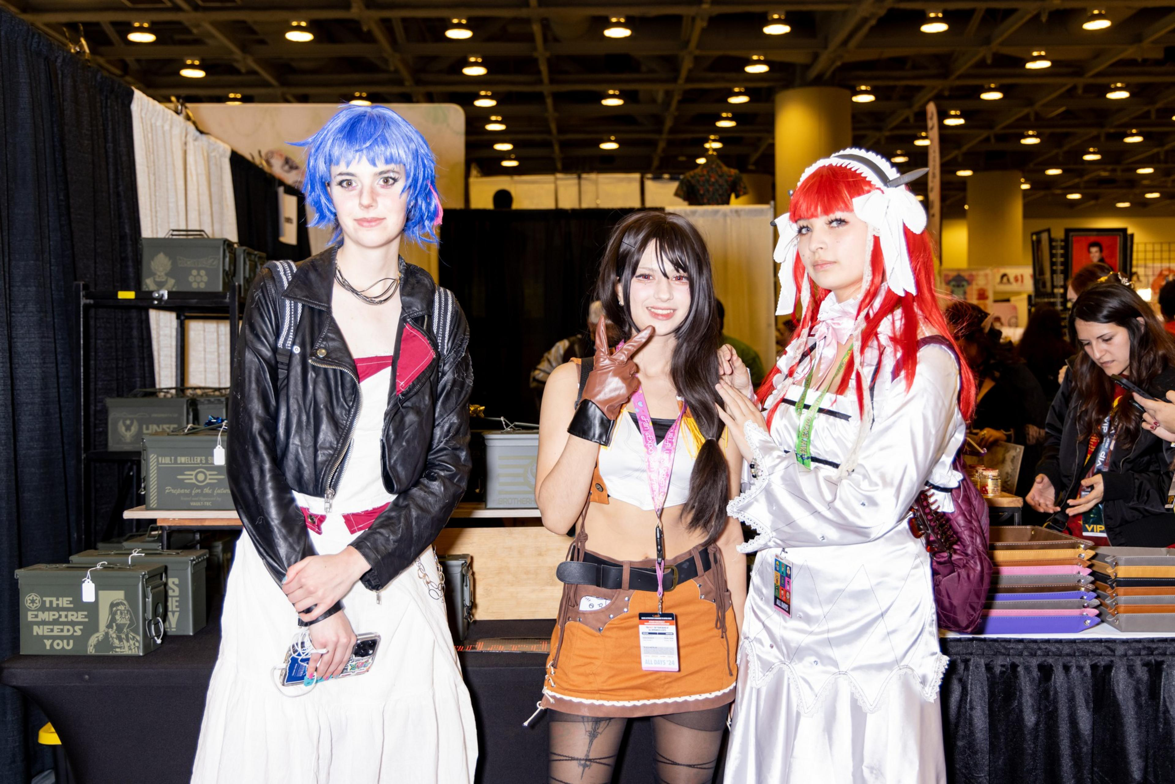 Three people are dressed in colorful costumes: one in blue hair with a black jacket, another with long brown hair and an orange outfit, and the third in red hair with a white dress, standing together indoors at what appears to be a convention.