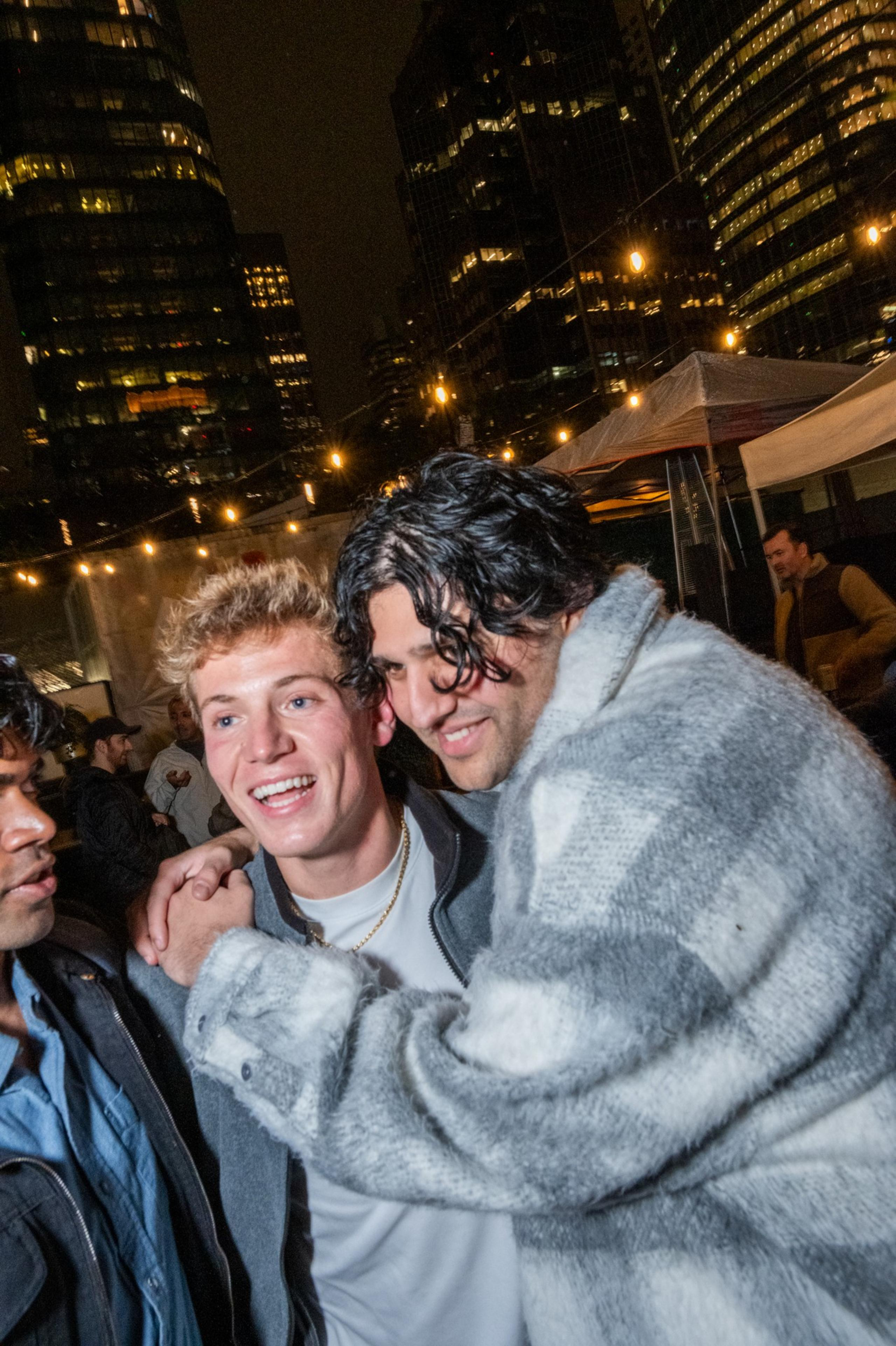 Three men are happily embracing at a nighttime outdoor gathering with city skyscrapers and string lights in the background.