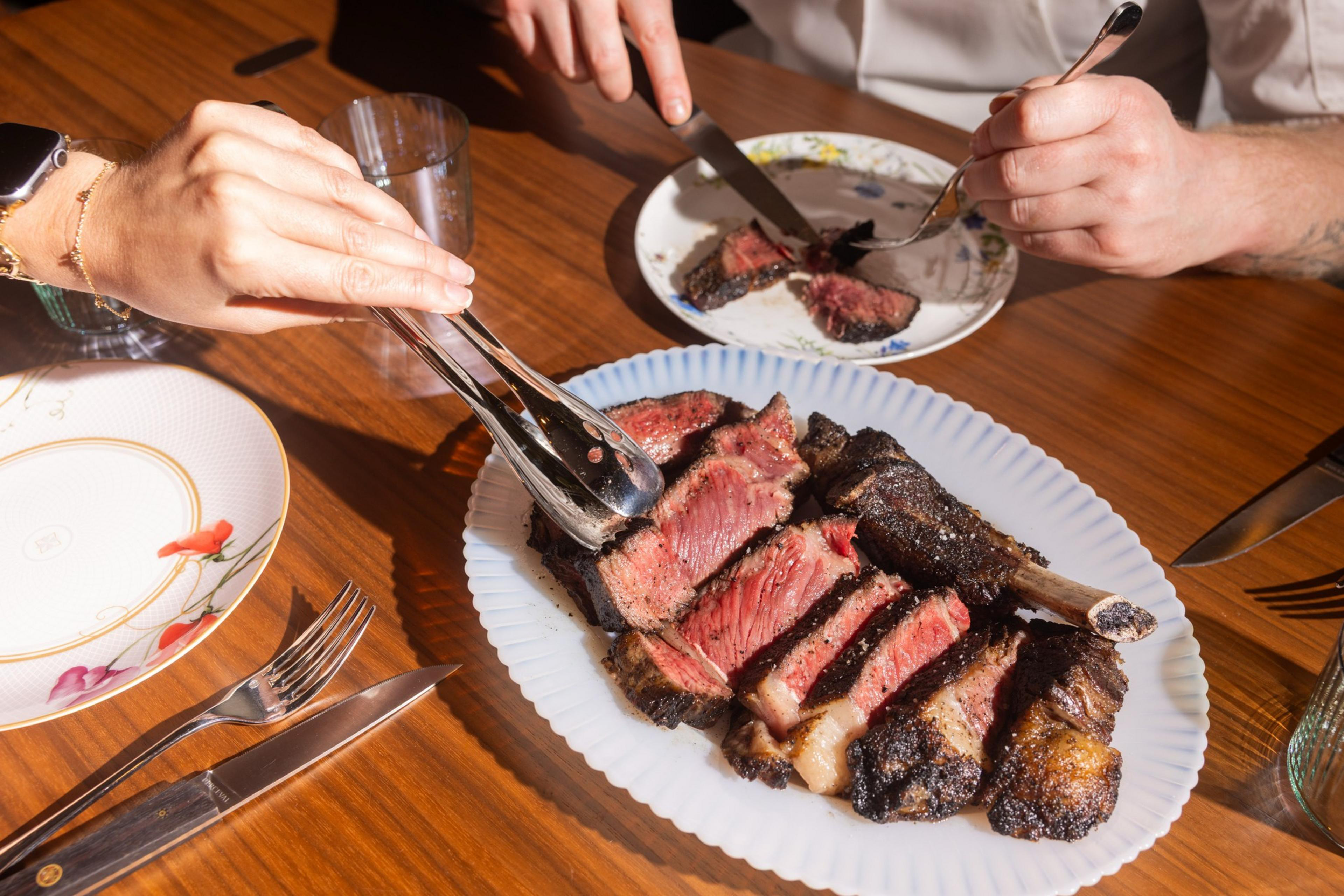 A wooden table with a large plate of sliced, medium-rare steak. Two people use utensils, with decorative plates, glasses, and cutlery nearby.