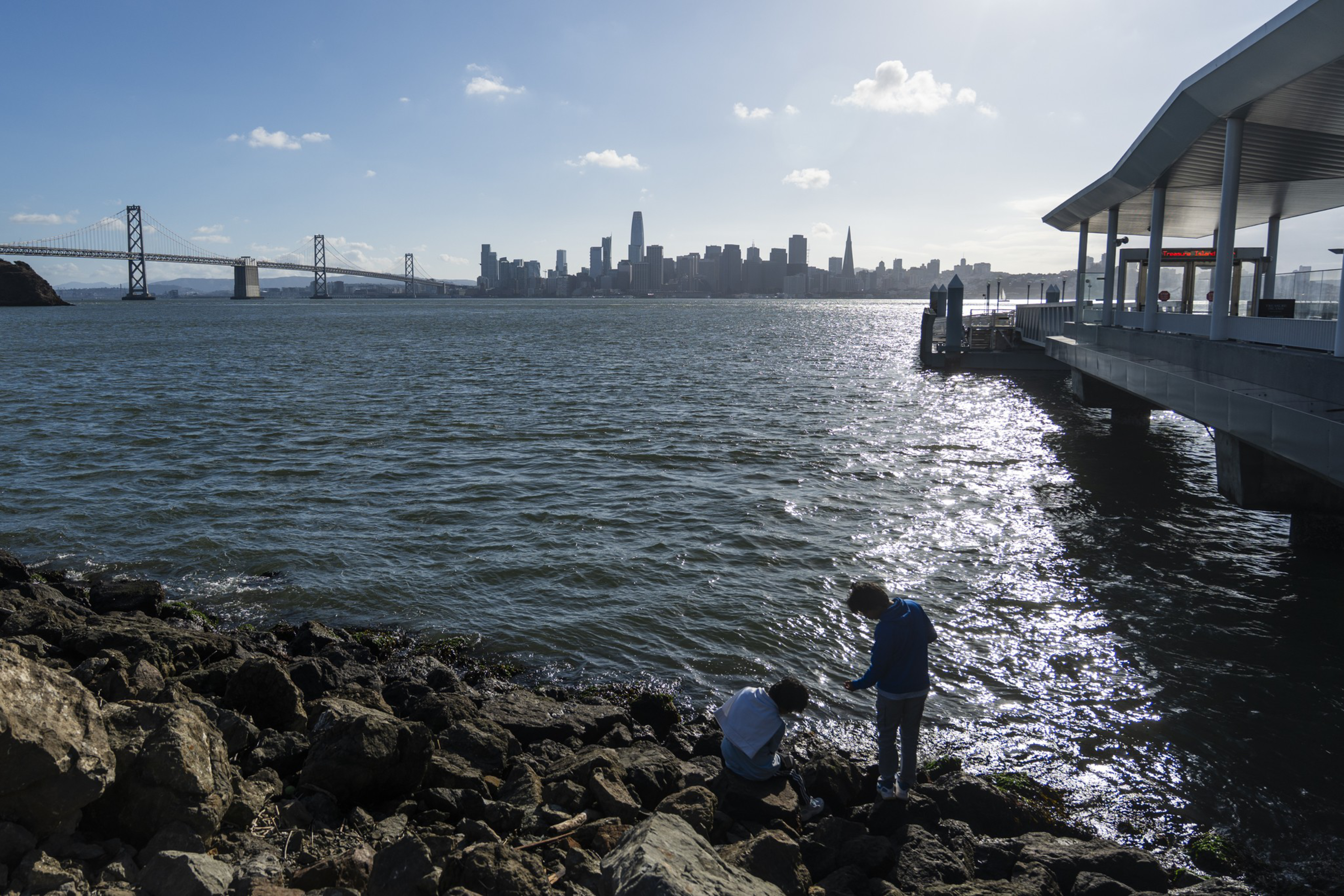 Two people are by rocky shore waters with a city skyline and bridge in the background, under a clear sky with sunlight reflecting on the water.