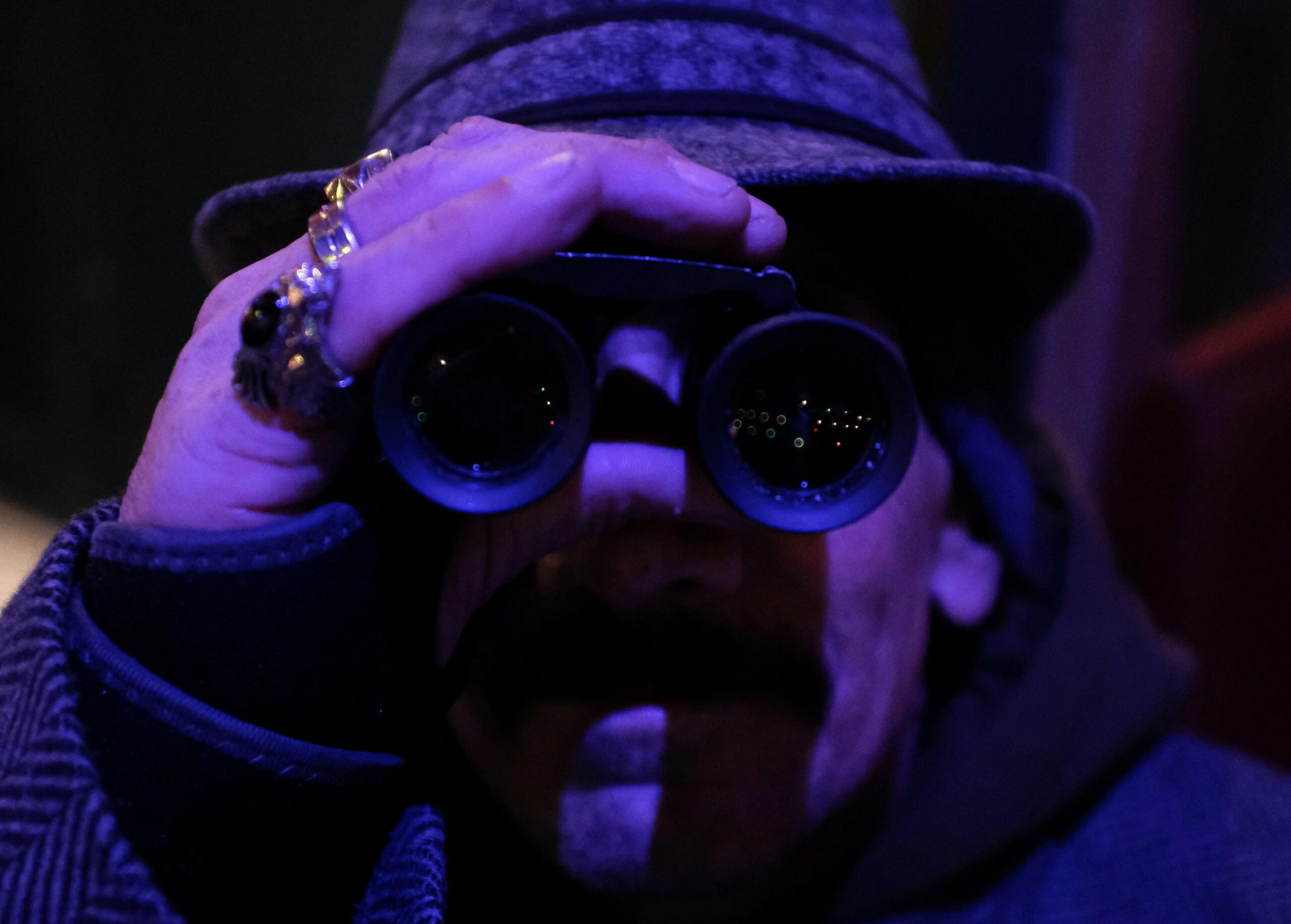 A person wearing a hat and multiple rings peers through binoculars in dim, moody blue-purple lighting.