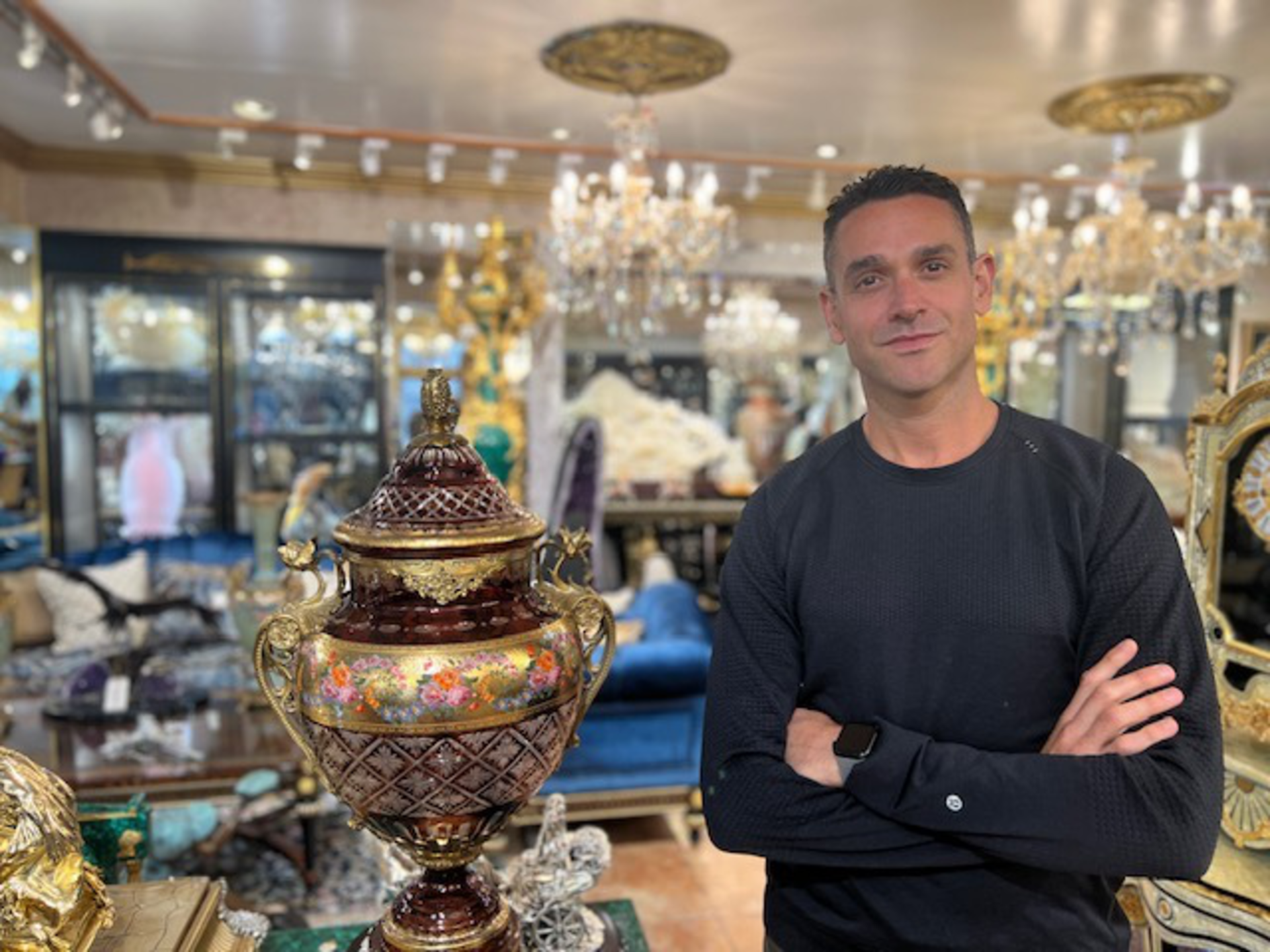 A man stands with his arms crossed surrounded by fine art objects.