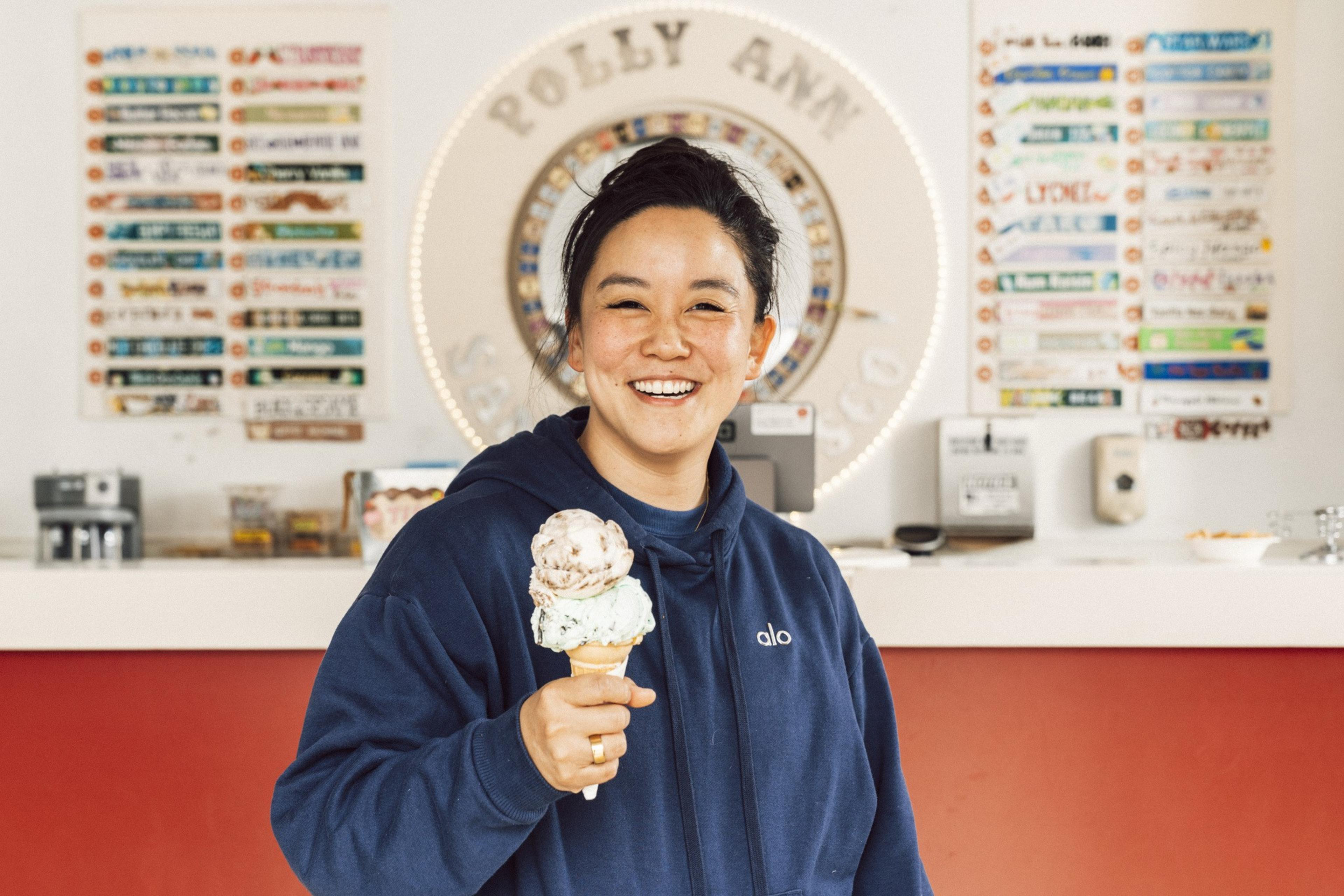 A person in a blue hoodie is smiling while holding an ice cream cone with two scoops. They're standing in front of a counter with colorful menus and signage.