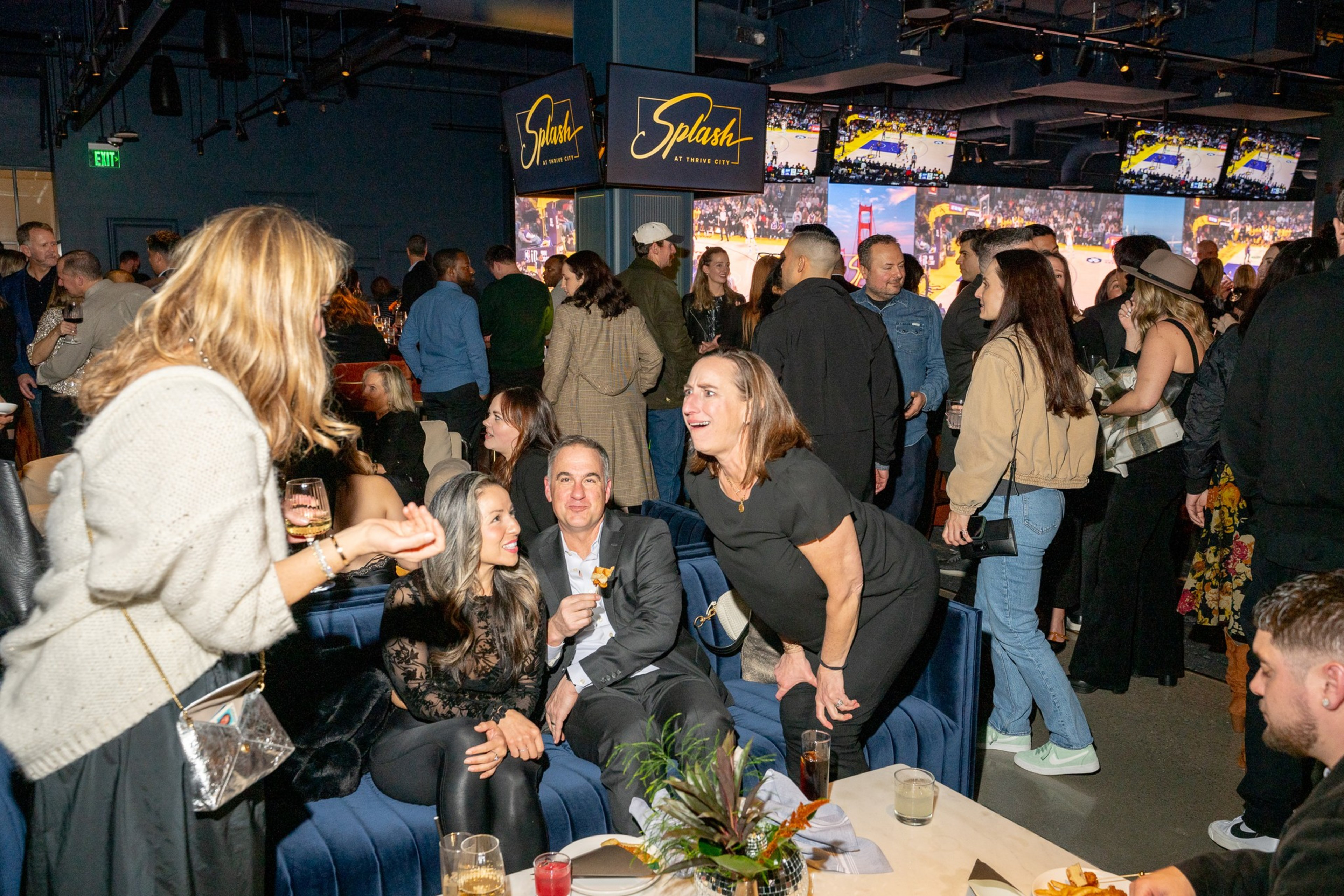 A lively indoor gathering with people socializing around sofas. TV screens show a basketball game. The atmosphere is vibrant with casual chatter.