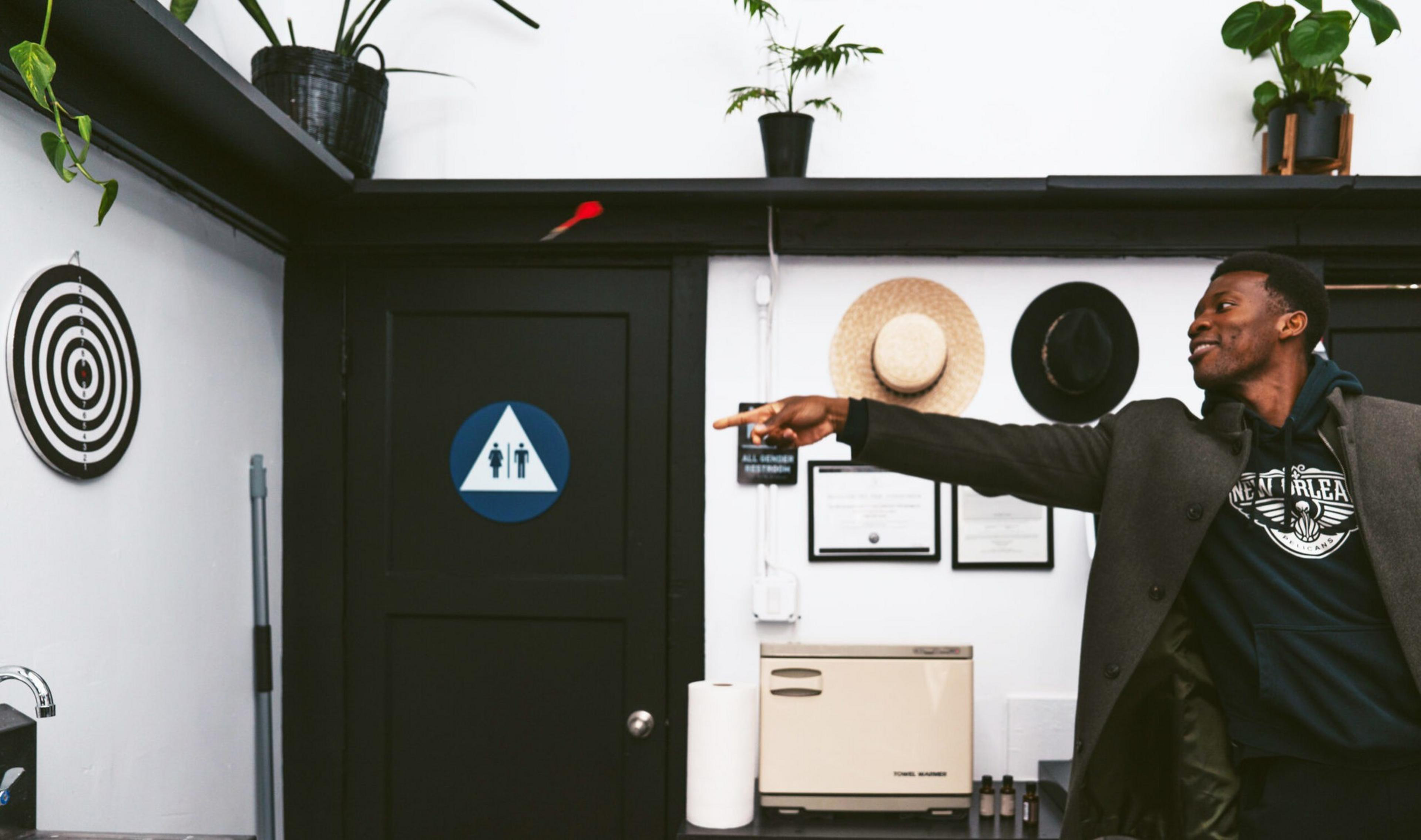 A man is throwing a red dart at a dartboard mounted on a black door with a restroom sign, surrounded by hats, plants, and framed documents.