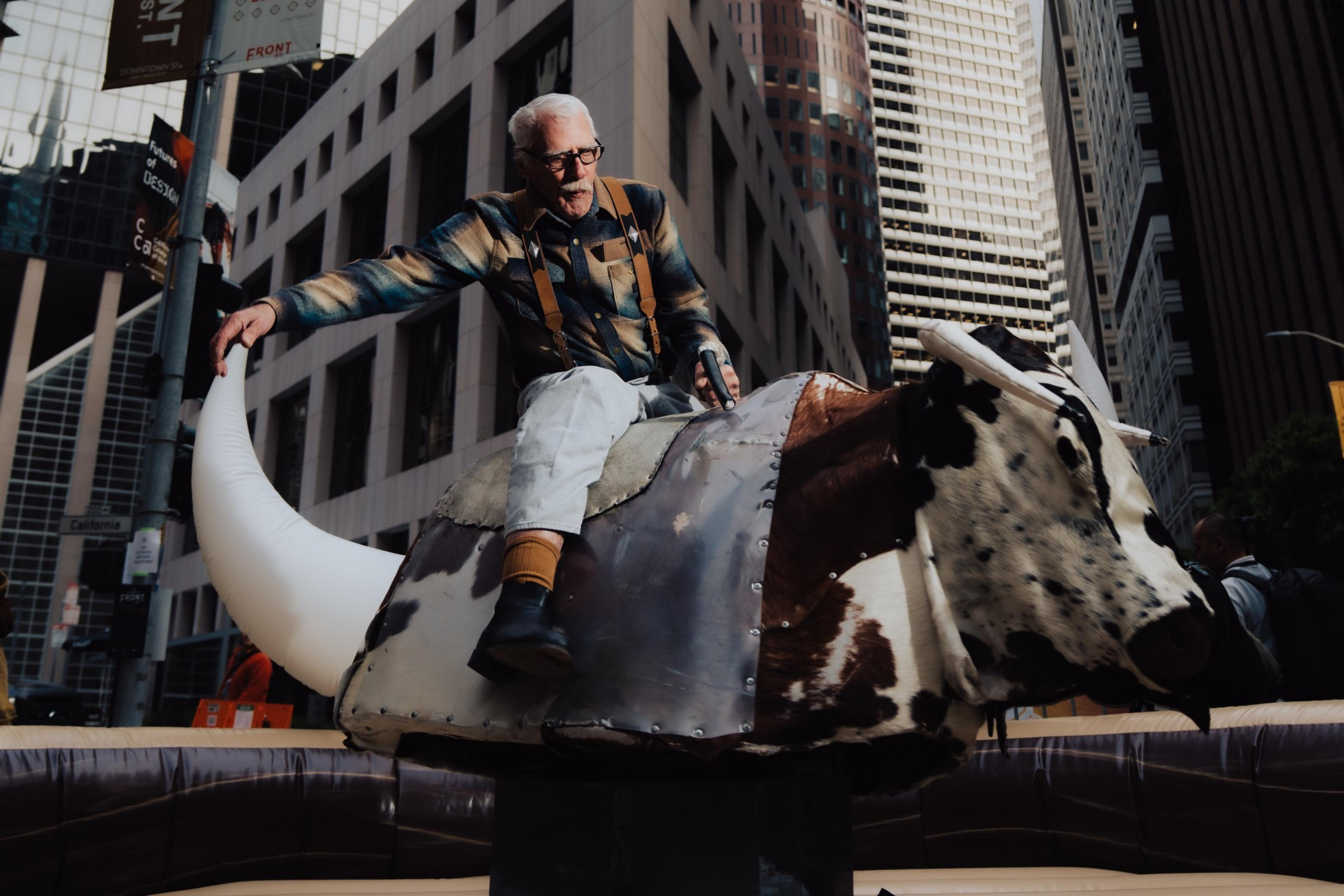 A man with white hair and glasses rides a mechanical bull in an urban setting, surrounded by tall buildings. The bull is brown and white with large horns.