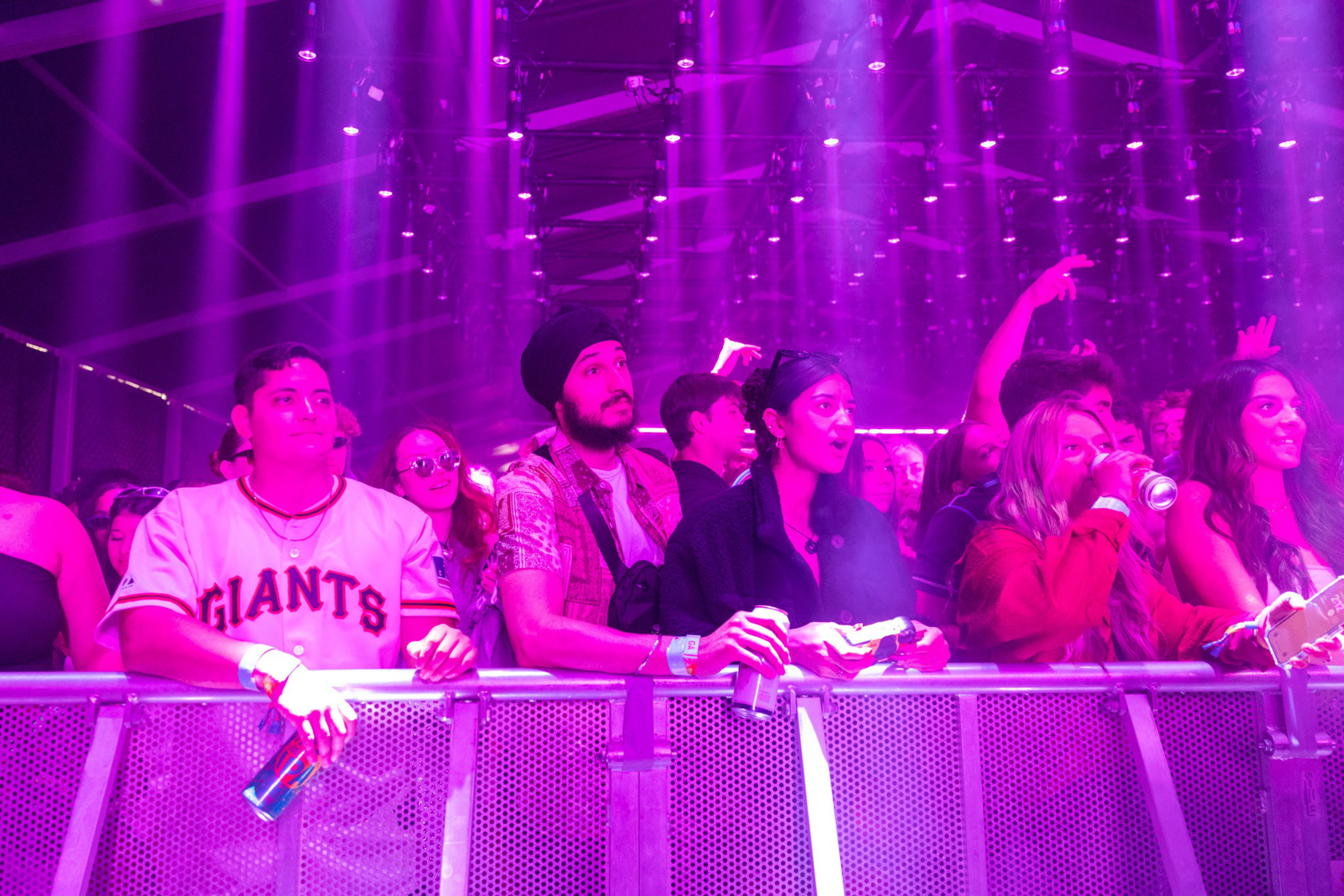 A diverse crowd of people stands at a barrier under purple stage lights, watching a performance with some holding drinks and wearing wristbands.