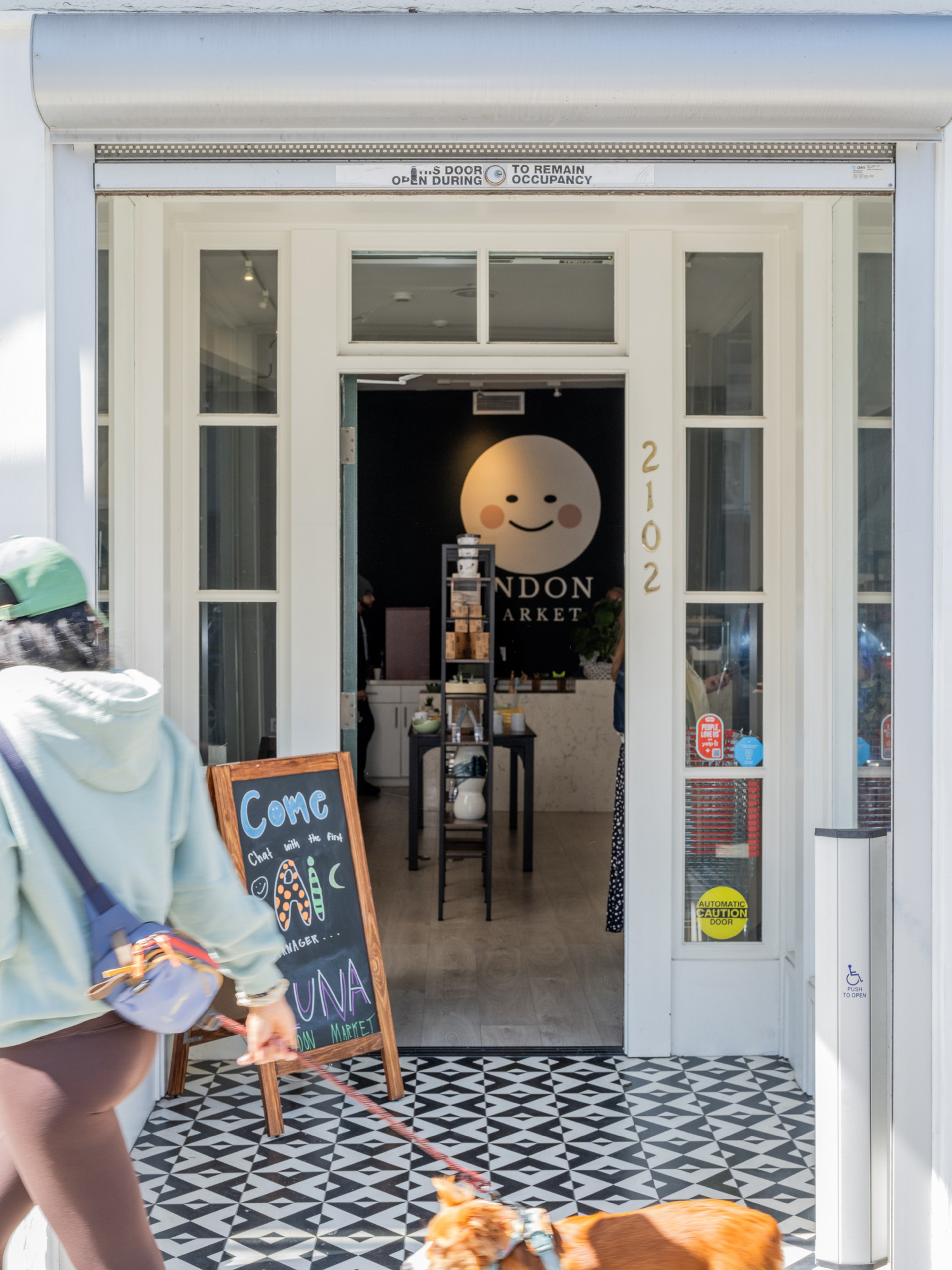 A person in a green cap walks a small dog past the open entrance of a market with a smiley face logo and a chalkboard sign outside.