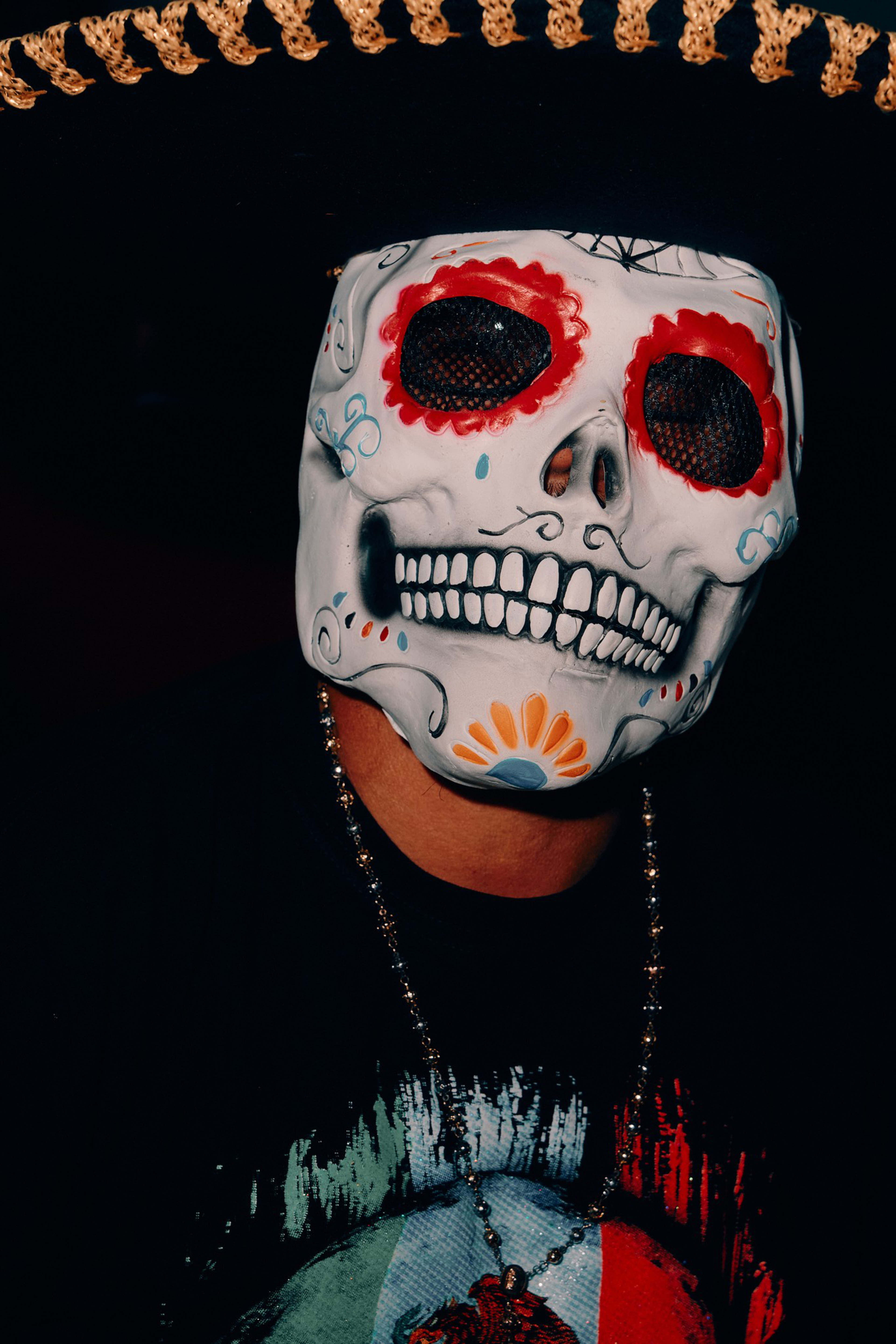 A person wears a white skull mask decorated with red, orange, and blue designs, along with a large black sombrero with brown trim.