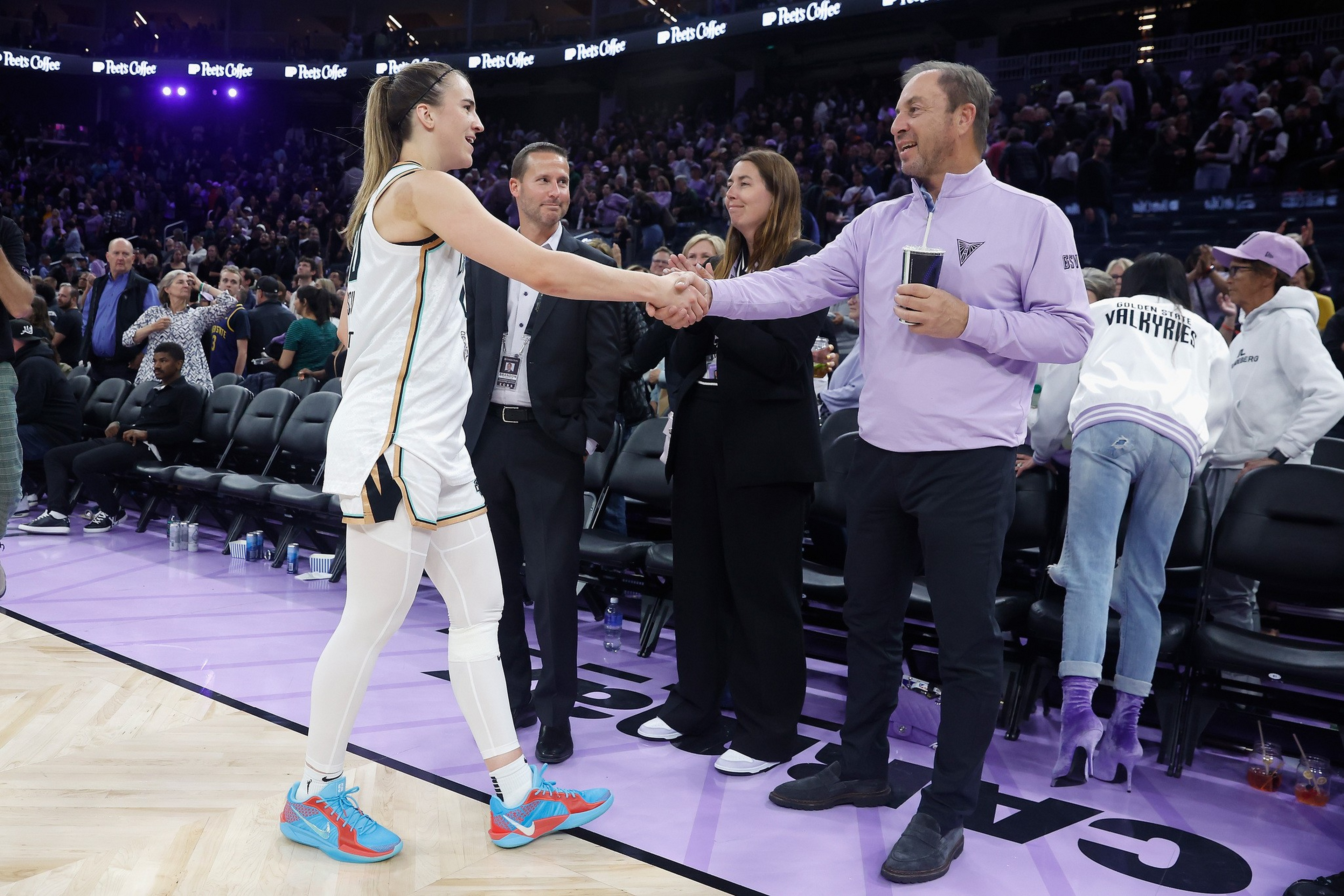 A basketball player in white shakes hands with a man in a lavender jacket courtside as a crowd watches in the background.