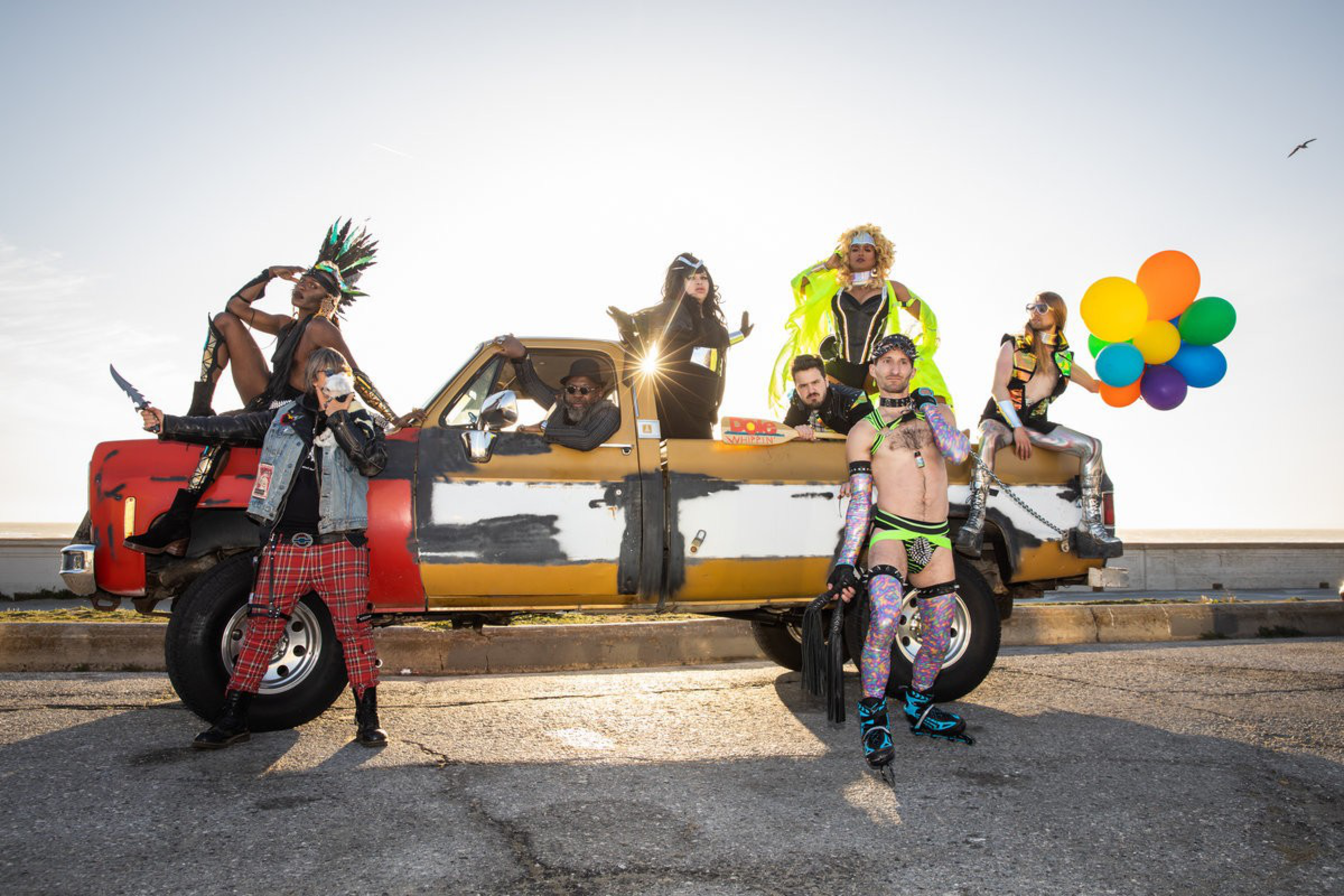 Seven people in colorful, eclectic outfits pose around and on a weathered pickup truck, with one person holding a cluster of vibrant balloons.